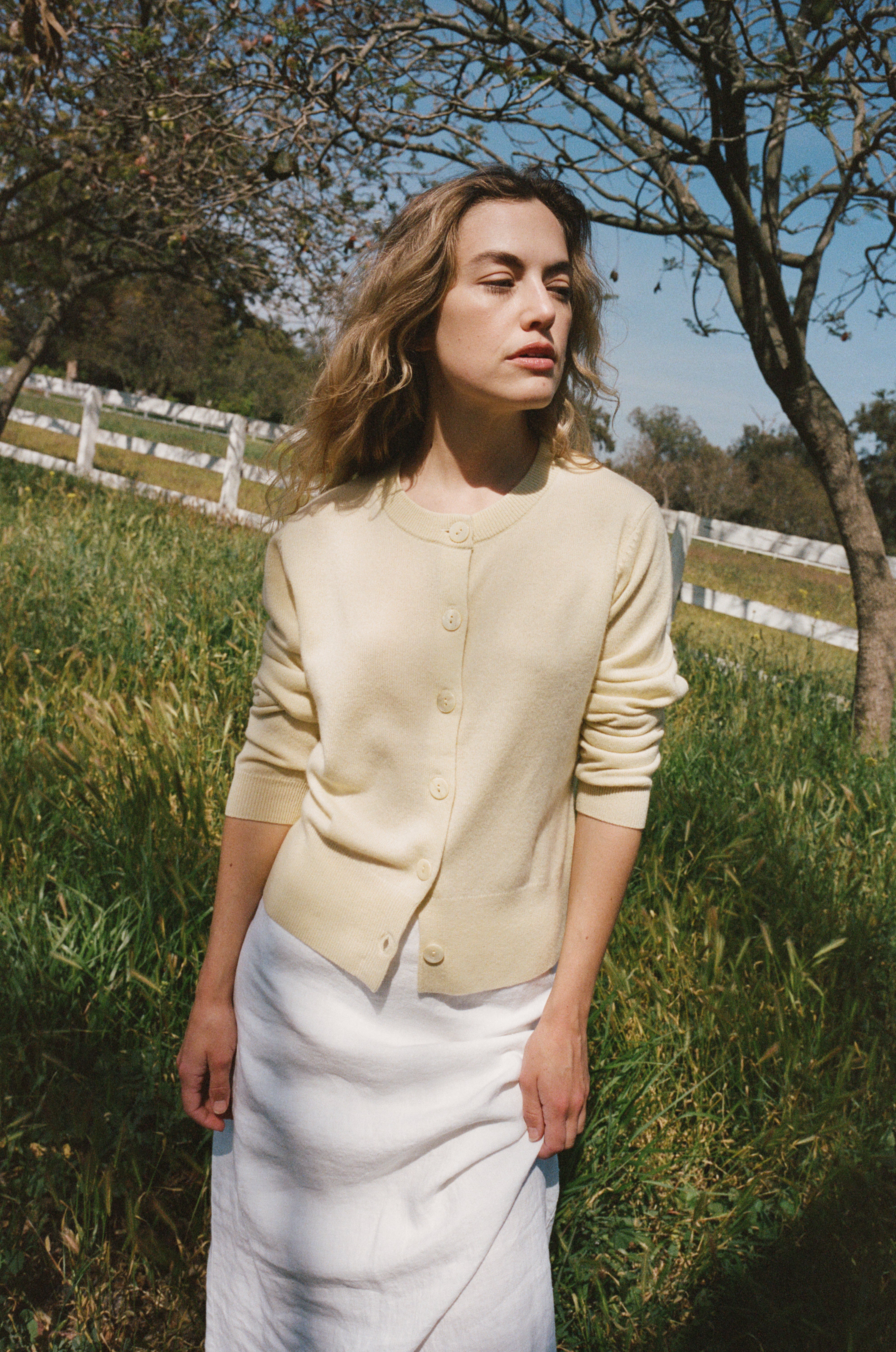 Female model leaning against a fence wearing a pale yellow ultra-soft luxurious cashmere button up cardigan paired with a white linen maxi skirt, and white flats - VIR VITA
#color_butter
