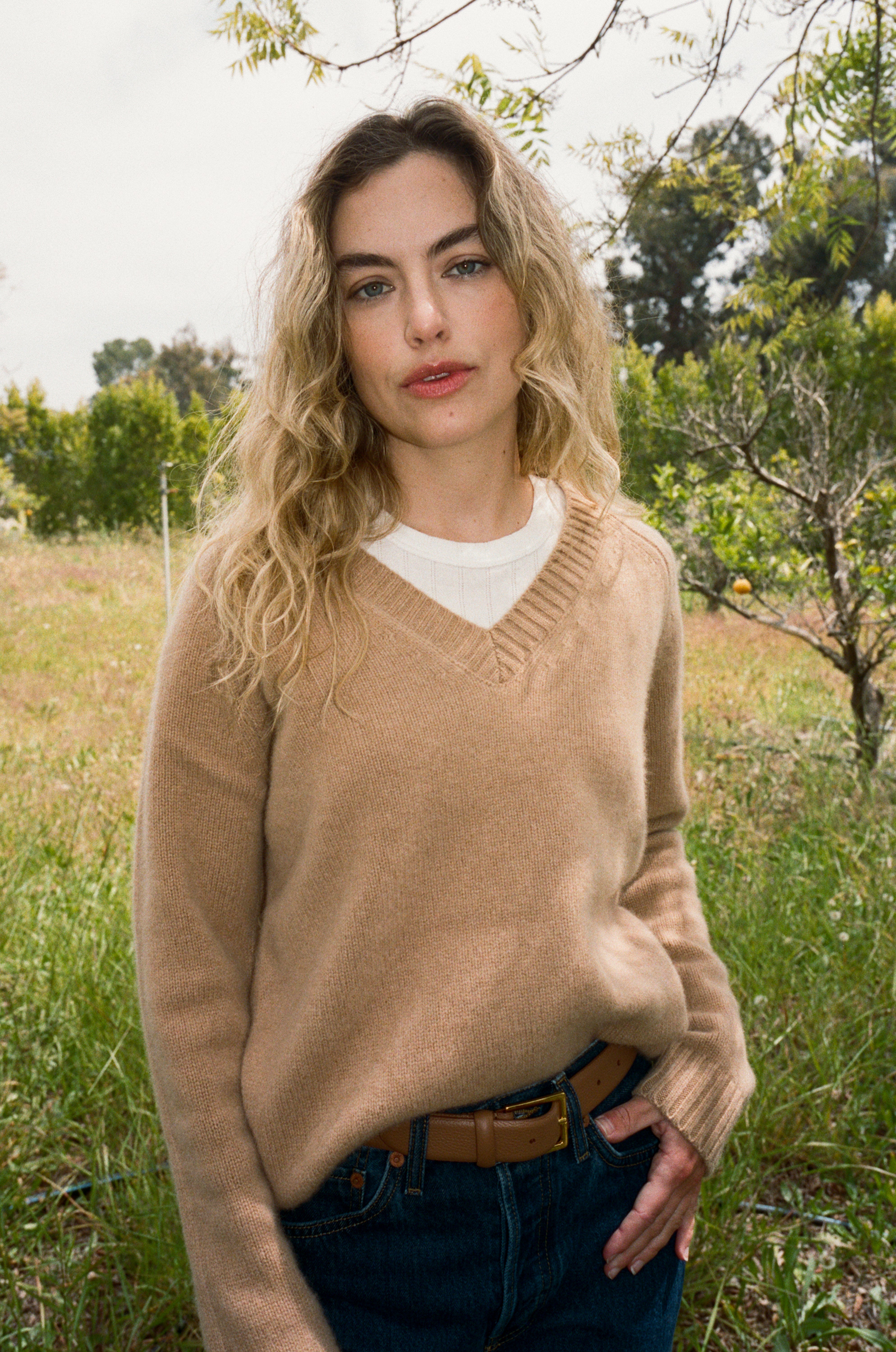 Female model standing in a field wearing an ultra-soft luxurious cashmere pullover sweater featuring a v-neck design in a light brown color paired with dark blue jeans, a brown belt, and a white pointelle tee - VIR VITA
#color_desert 
