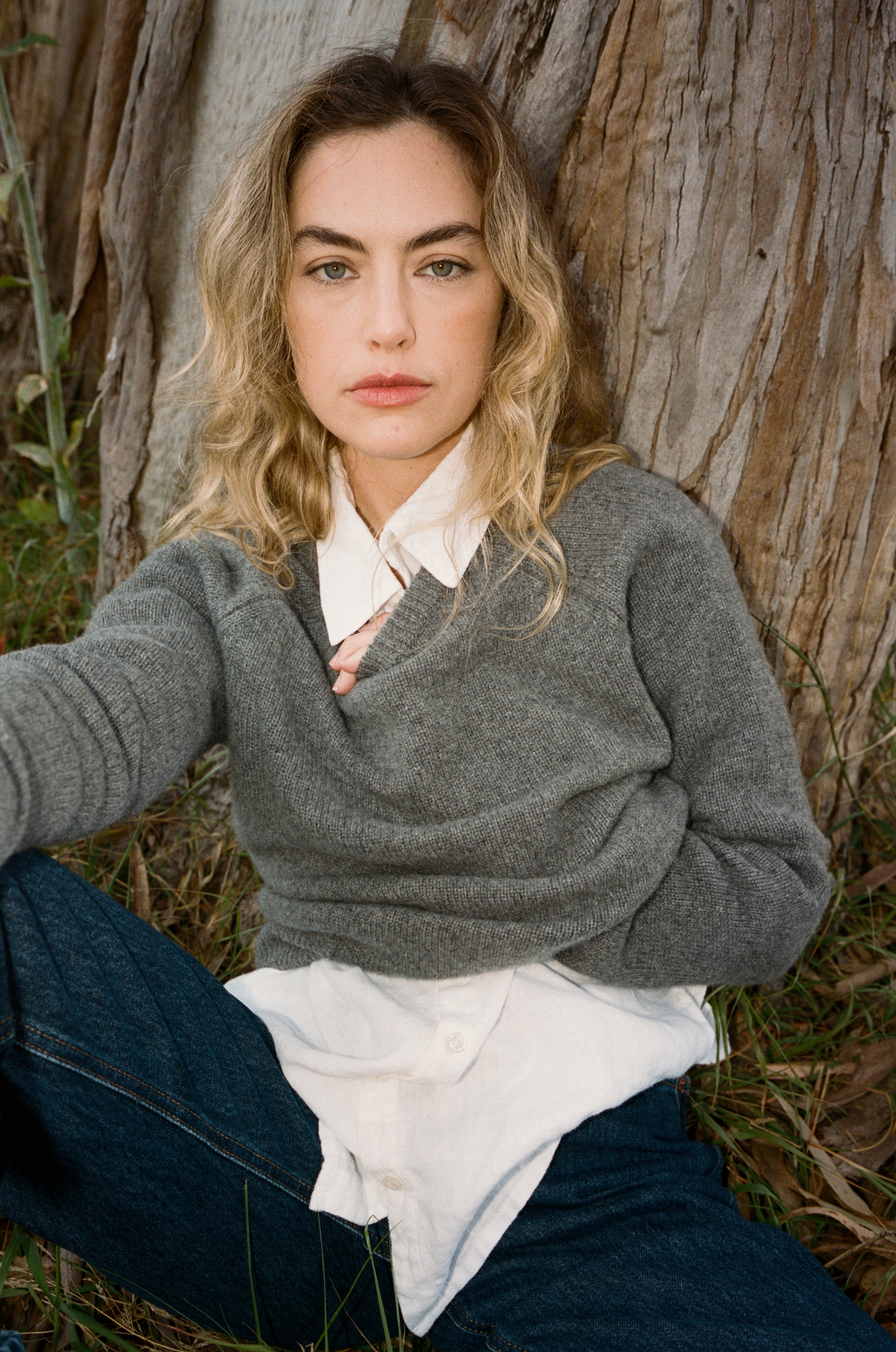 Female model sitting next to a tree wearing a grey ultra-soft luxurious cashmere pullover sweater featuring a classic silhouette and raglan sleeves paired with a white collared shirt and blue jeans - VIR VITA
#color_stone grey
