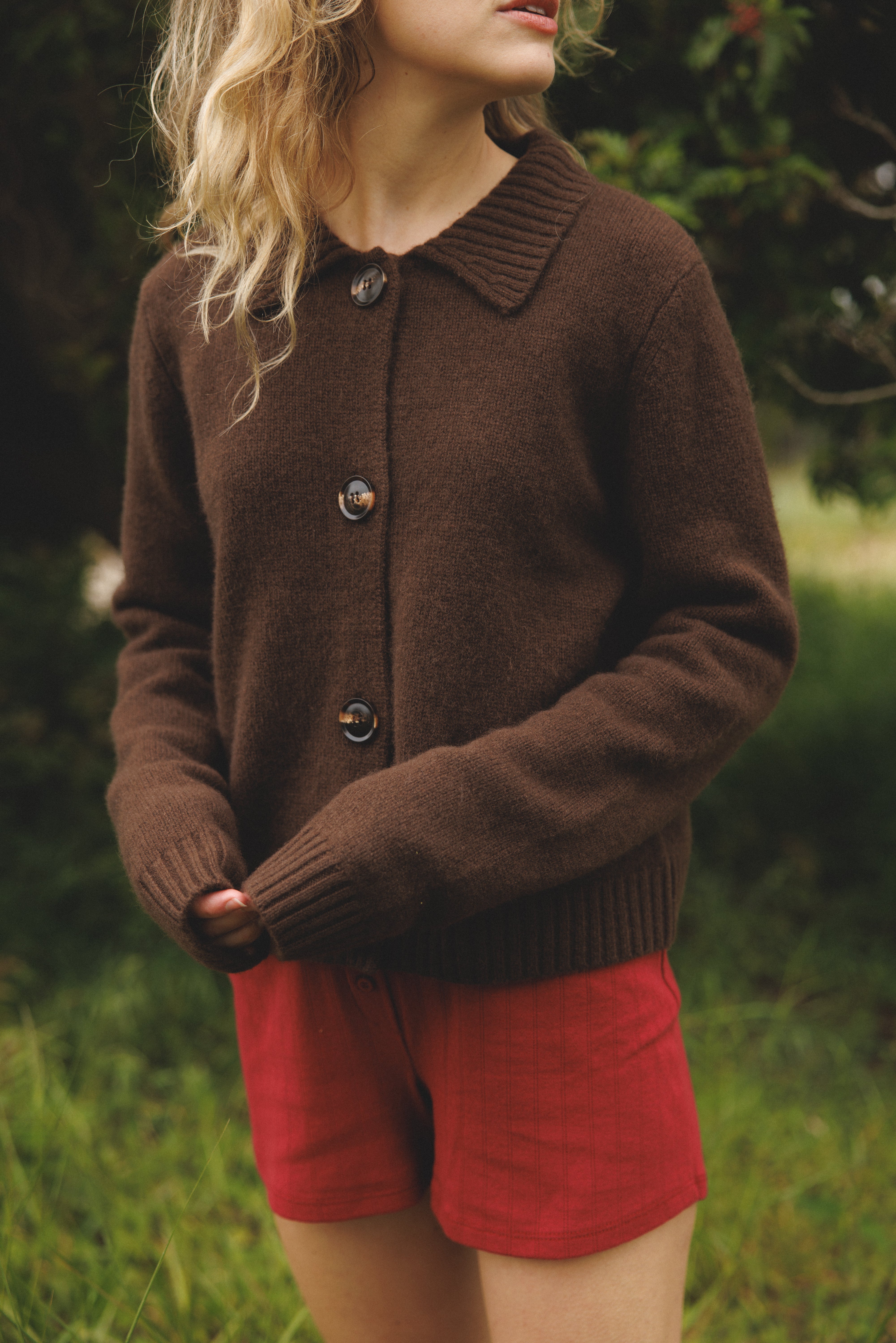 Female model standing in grass wearing a dark brown ultra-soft luxurious cashmere cardigan featuring a classic silhouette with a collar and large buttons paired with red pointelle shorts - VIR VITA
#color_espresso