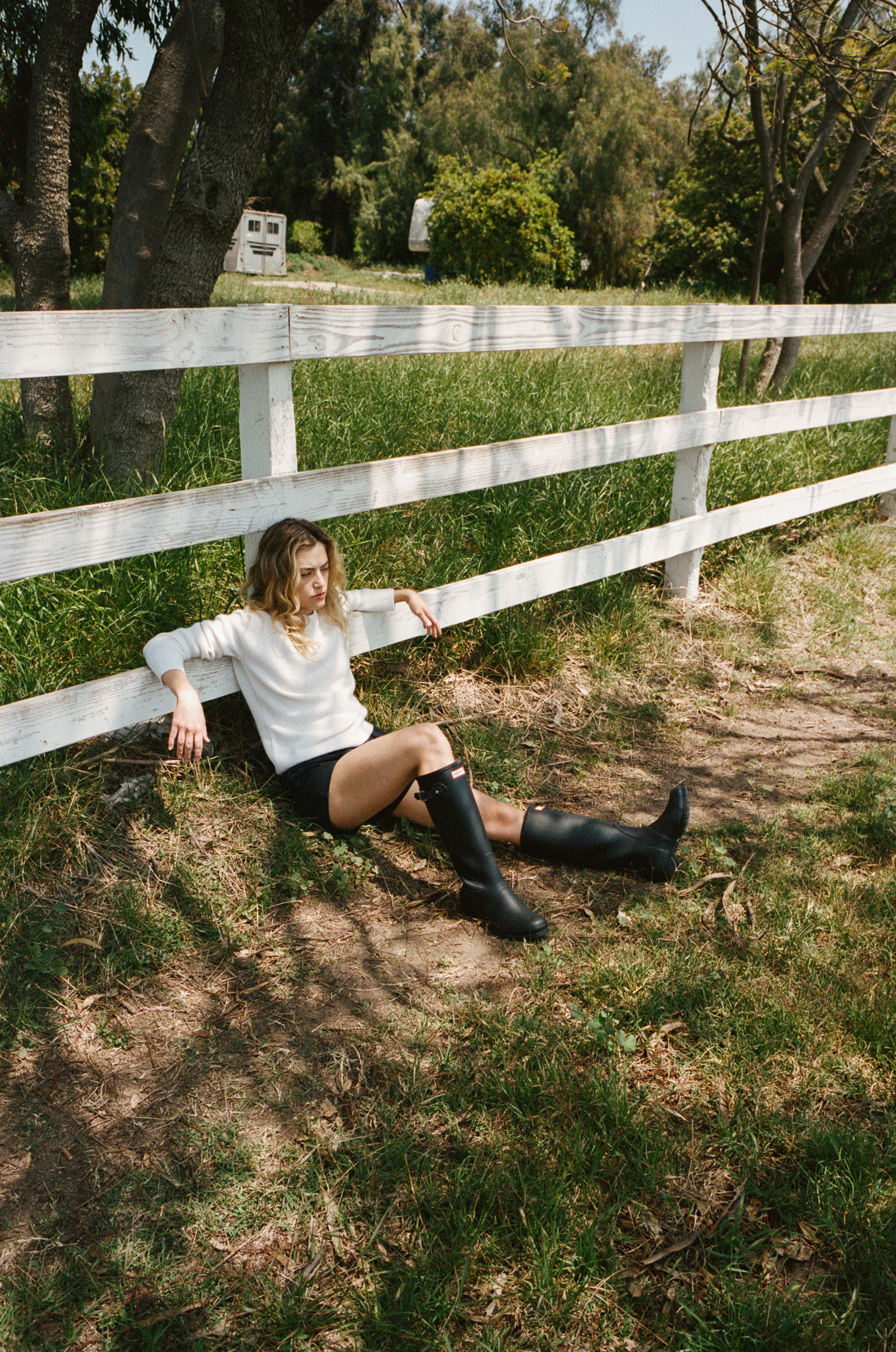 Female model leaning on a fence wearing a white ultra-soft luxurious cashmere pullover sweater featuring a classic silhouette and raglan sleeves paired with black cotton poplin shorts, and tall black boots - VIR VITA
#color_bone