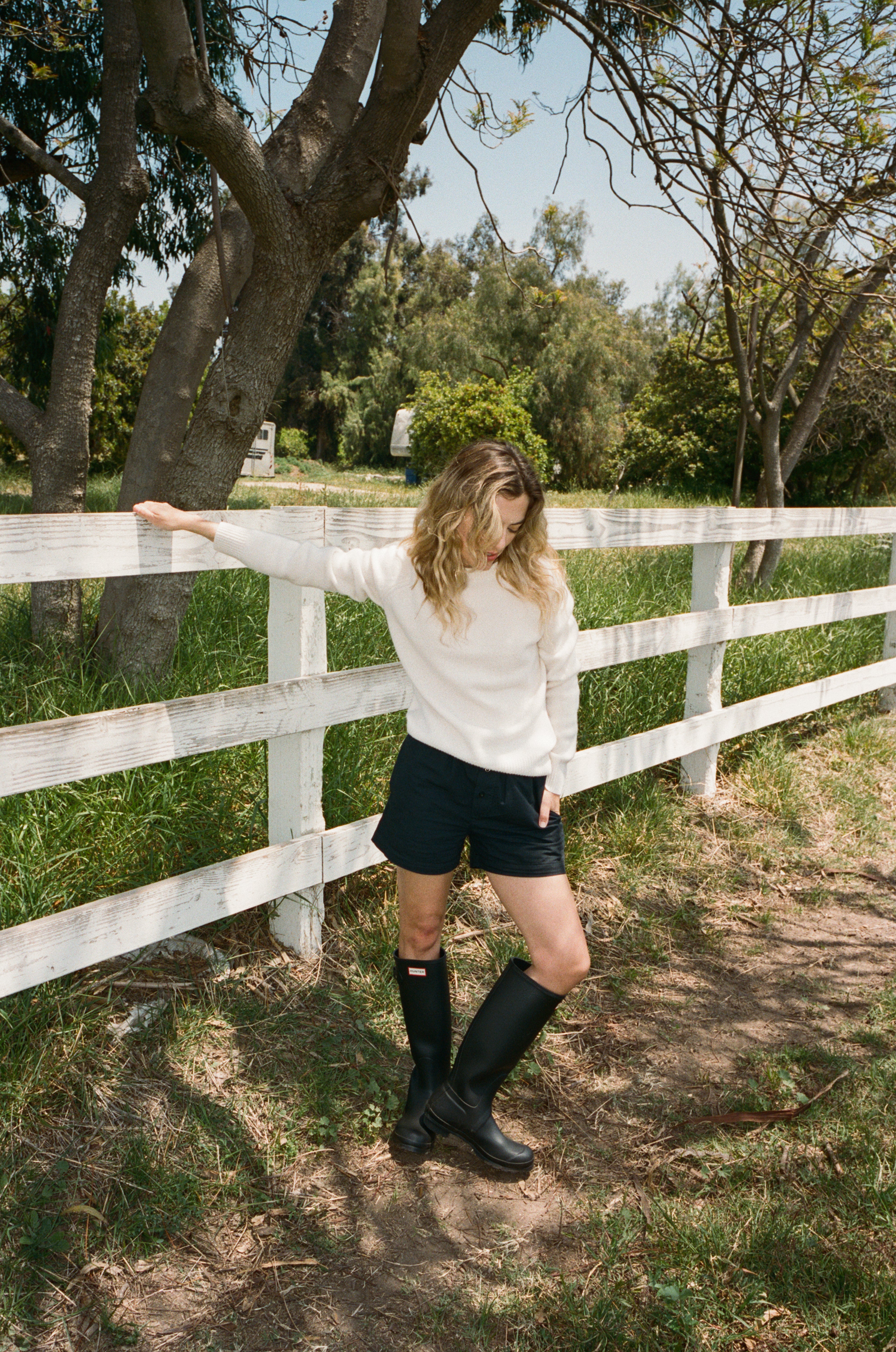 Female model standing next to a fence wearing a white ultra-soft luxurious cashmere pullover sweater featuring a classic silhouette and raglan sleeves paired with black cotton poplin shorts, and tall black boots - VIR VITA
#color_bone