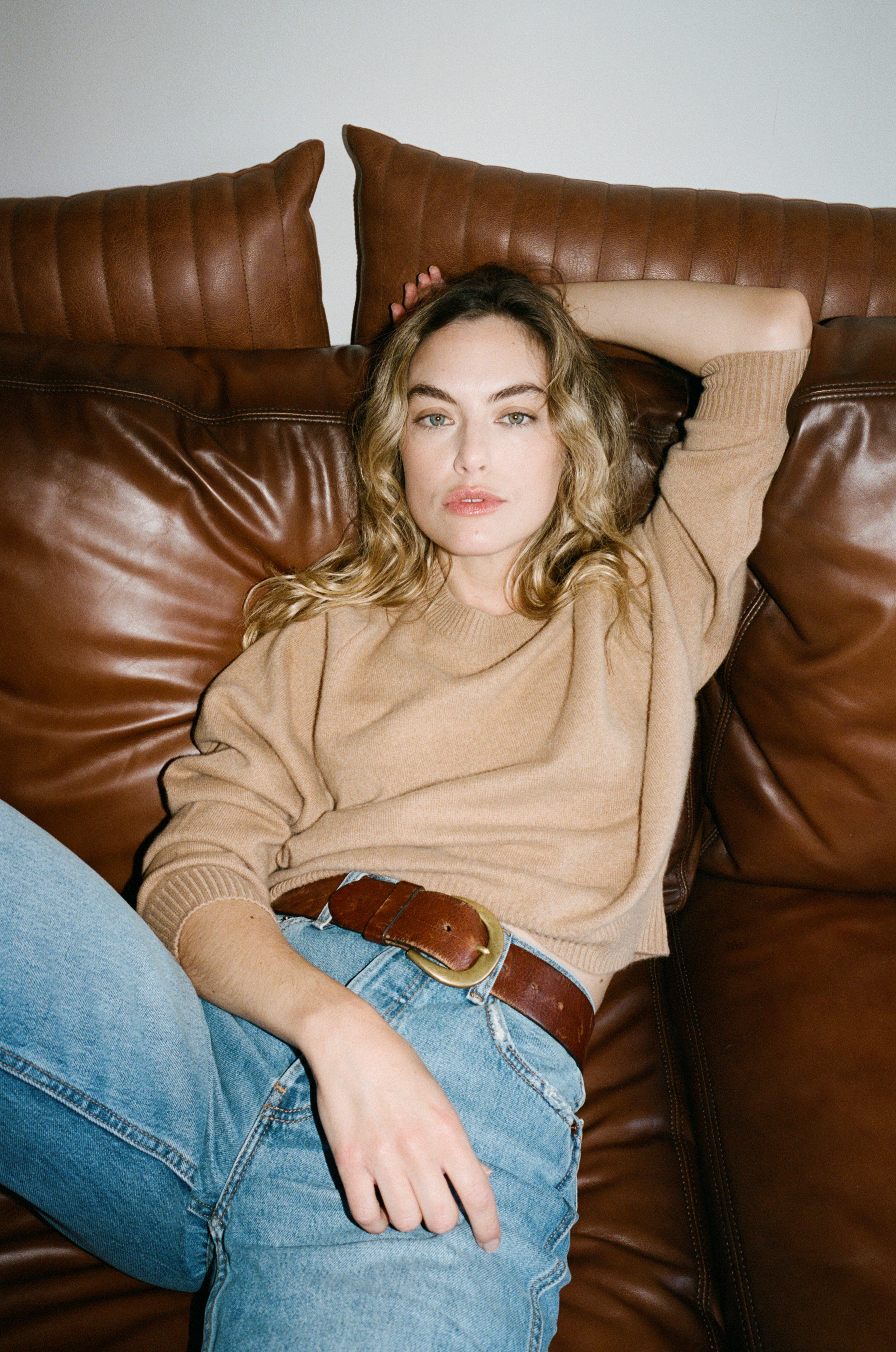 Female model laying on a couch wearing a  light brown luxurious and soft cashmere pullover sweater with a classic and relaxed silhouette paired with blue jeans and a brown belt - VIR VITA
#color_desert
