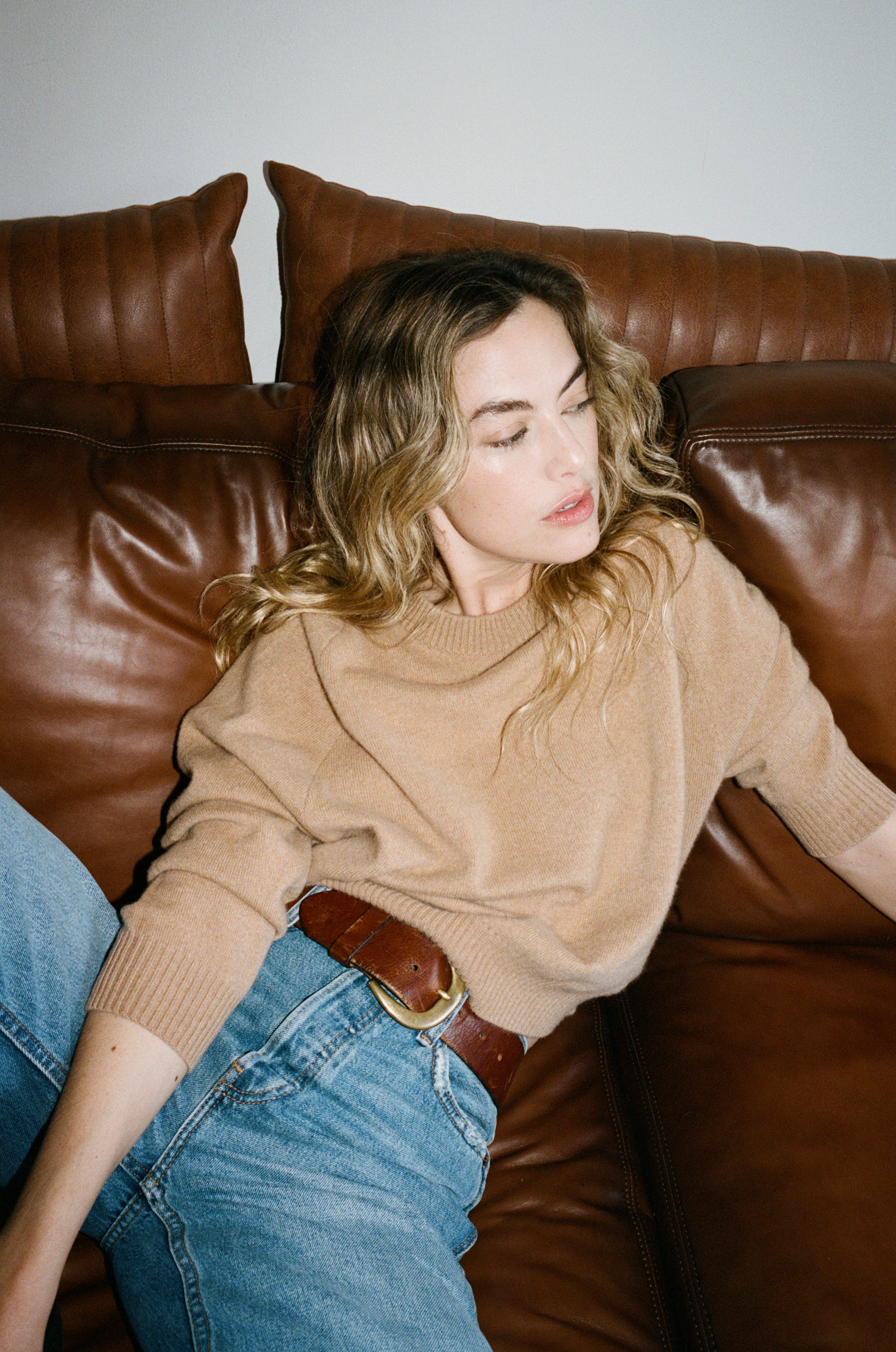Female model laying on a couch wearing a  light brown luxurious and soft cashmere pullover sweater with a classic and relaxed silhouette paired with blue jeans and a brown belt - VIR VITA
#color_desert