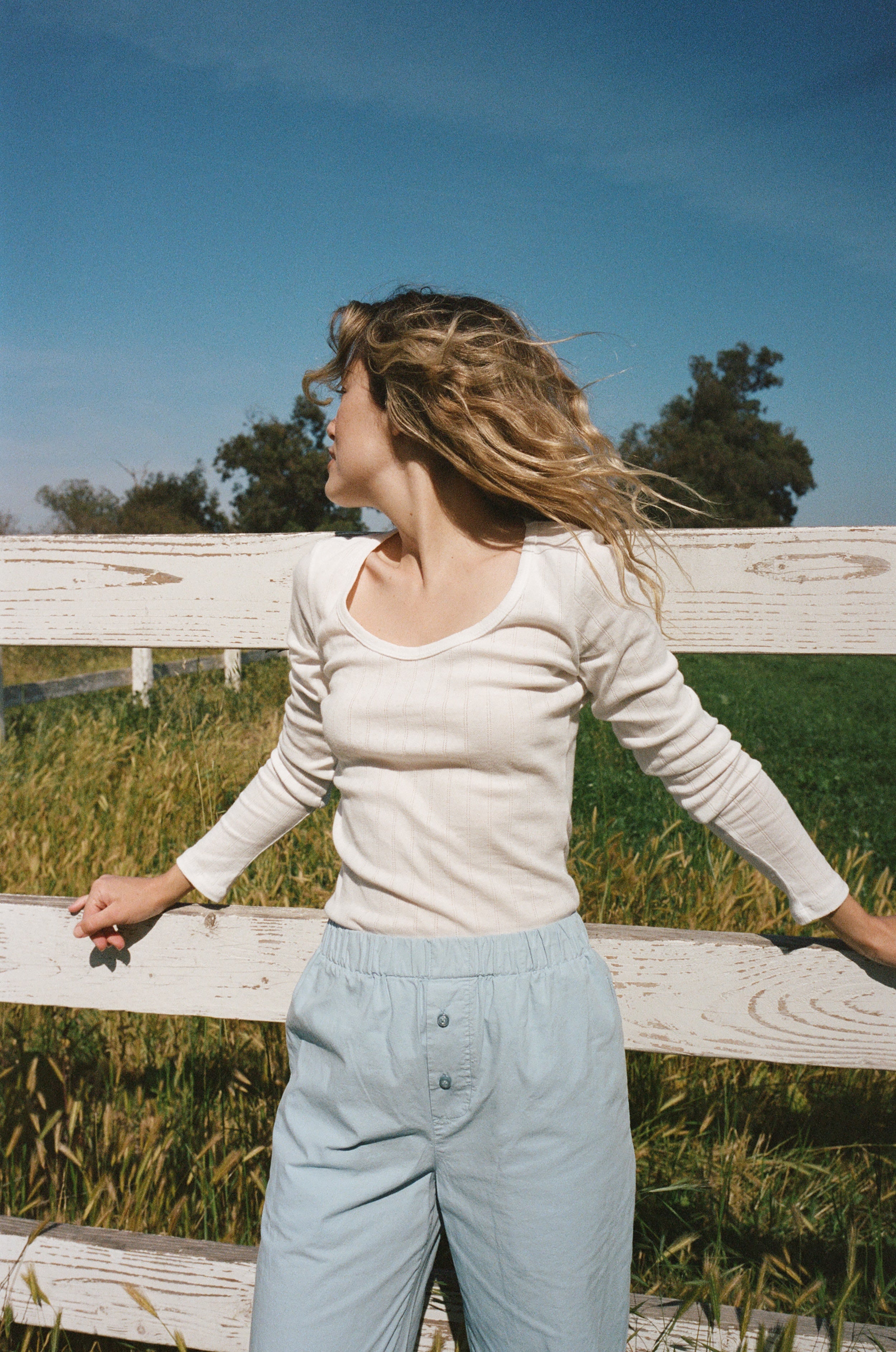 Female model leaning against a fence wearing a white pointelle long sleeve top featuring a flattering scoop neckline paired with cotton poplin pants in light blue - VIR VITA
#color_coconut