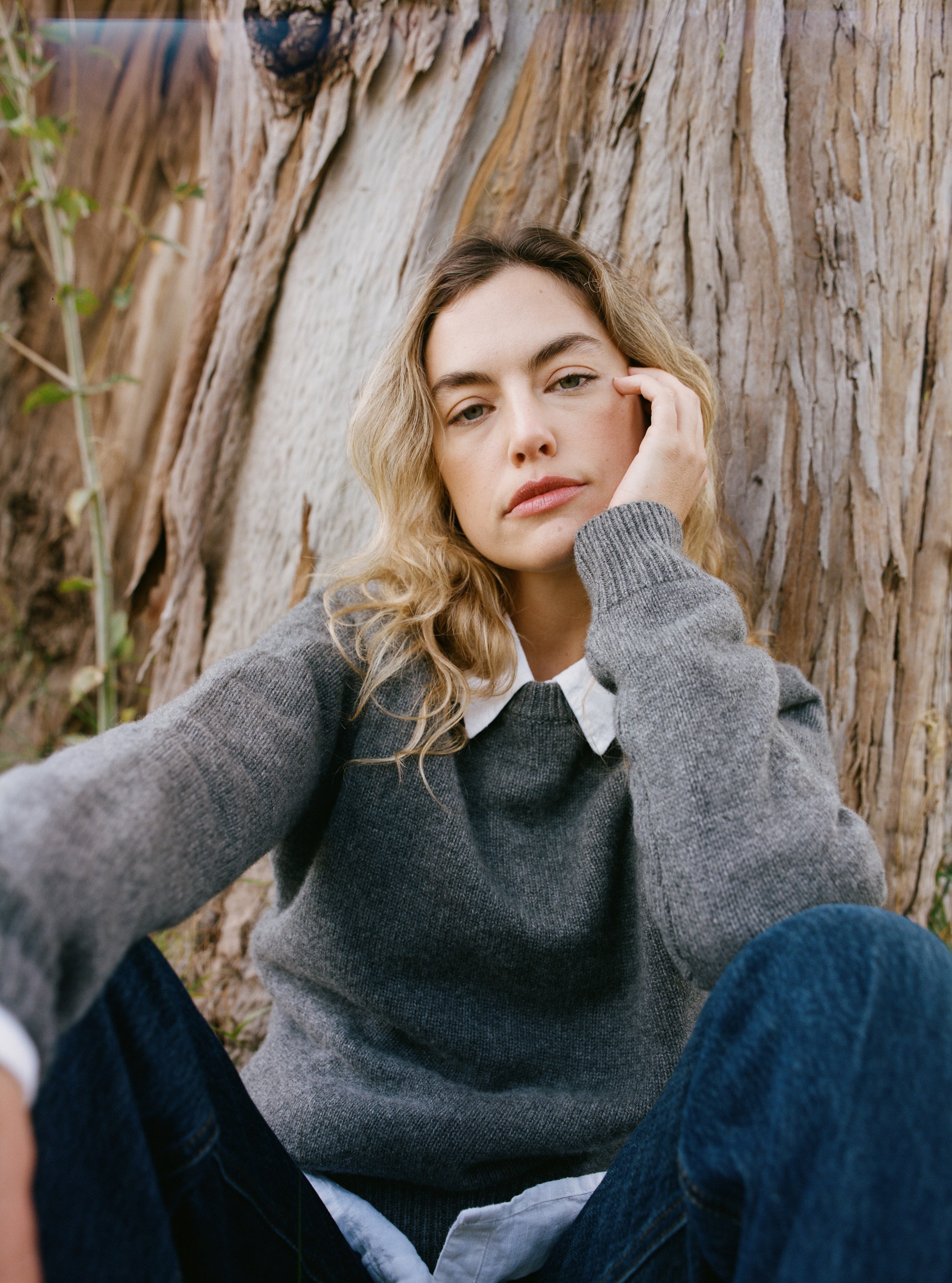 Female model sitting next to a tree wearing a grey ultra-soft luxurious cashmere pullover sweater featuring a classic silhouette and raglan sleeves paired with a white collared shirt and blue jeans - VIR VITA
#color_stone grey