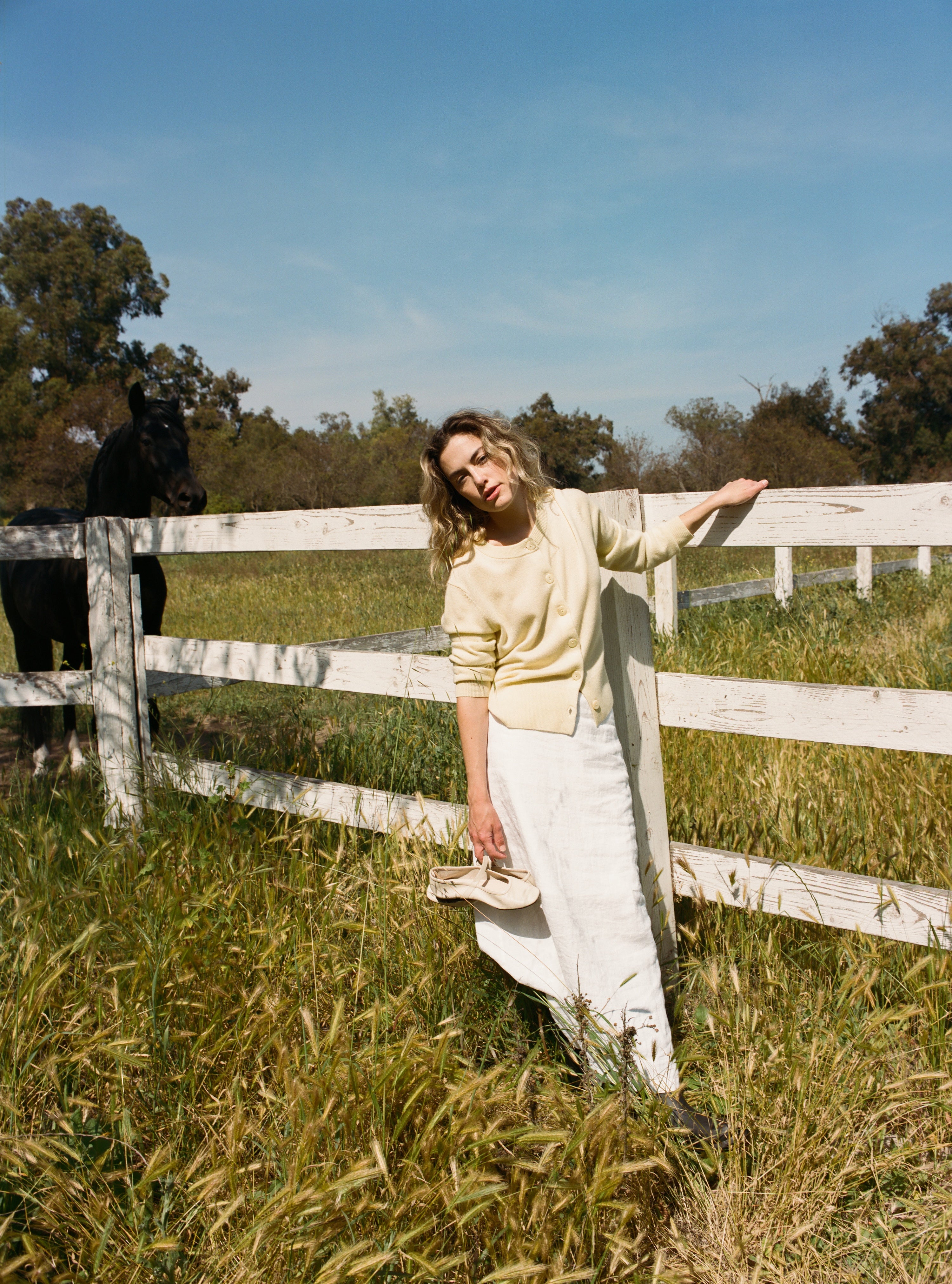 Female model leaning against a fence wearing a pale yellow ultra-soft luxurious cashmere button up cardigan paired with a white linen maxi skirt, and white flats - VIR VITA
#color_butter