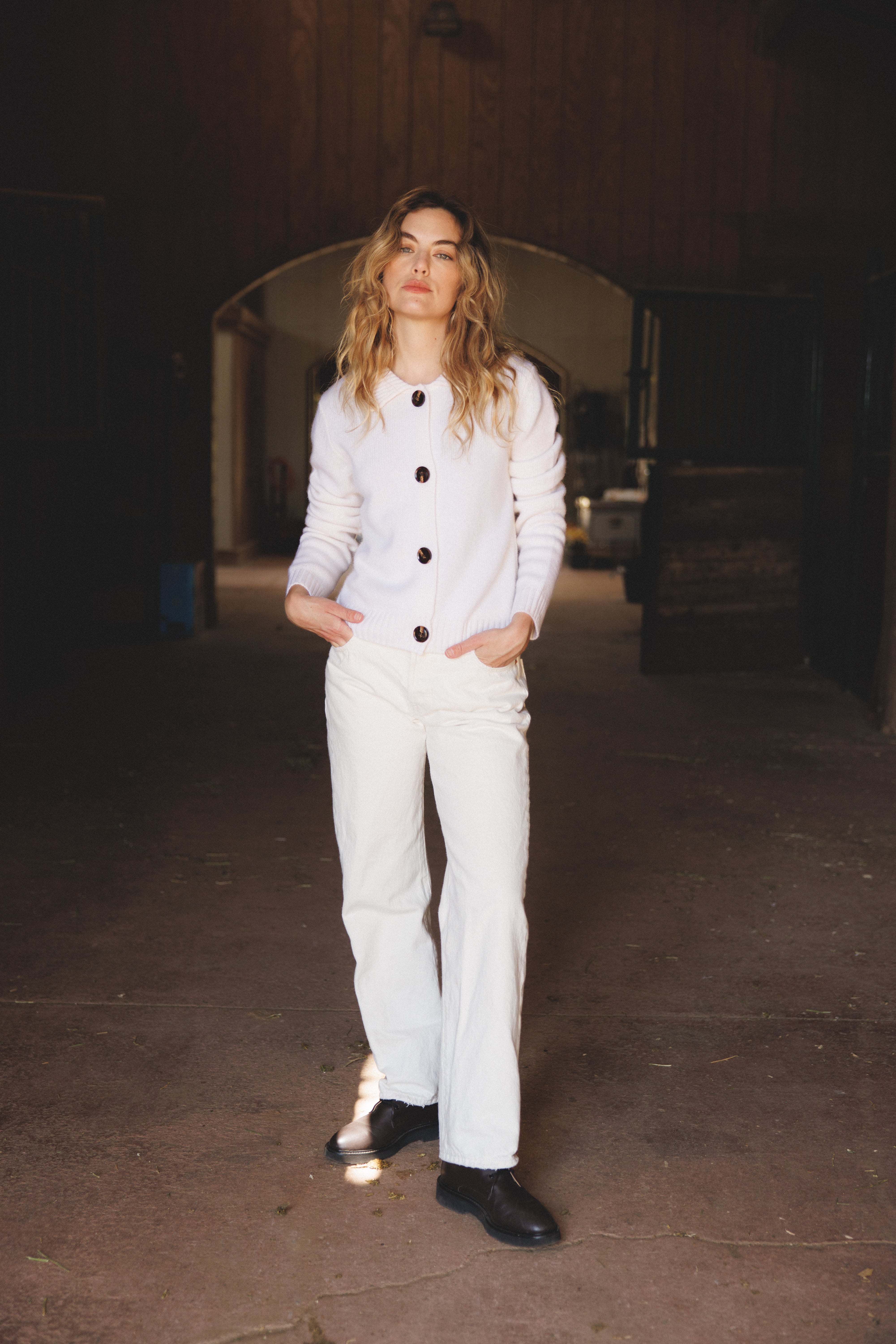 Female model wearing a white ultra-soft luxurious cashmere cardigan featuring a classic silhouette with a collar and large buttons paired with white jeans and brown boots - VIR VITA
#color_bone
