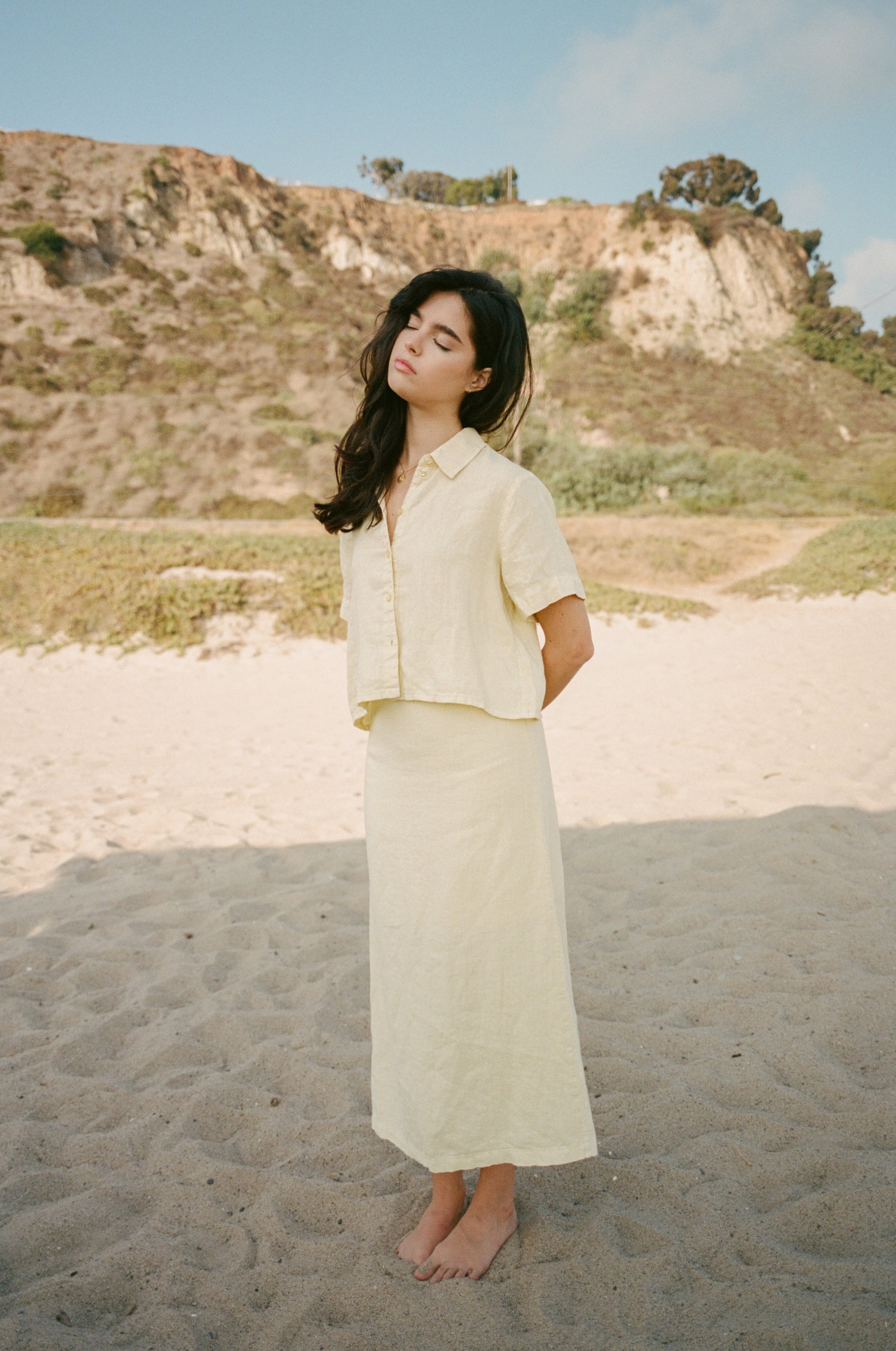 Female model wearing a premium 100% linen maxi skirt featuring a high waisted silhouette in pale yellow paired with a linen button up short sleeve shirt in pale yellow on the beach- VIR VITA 
#color_butter