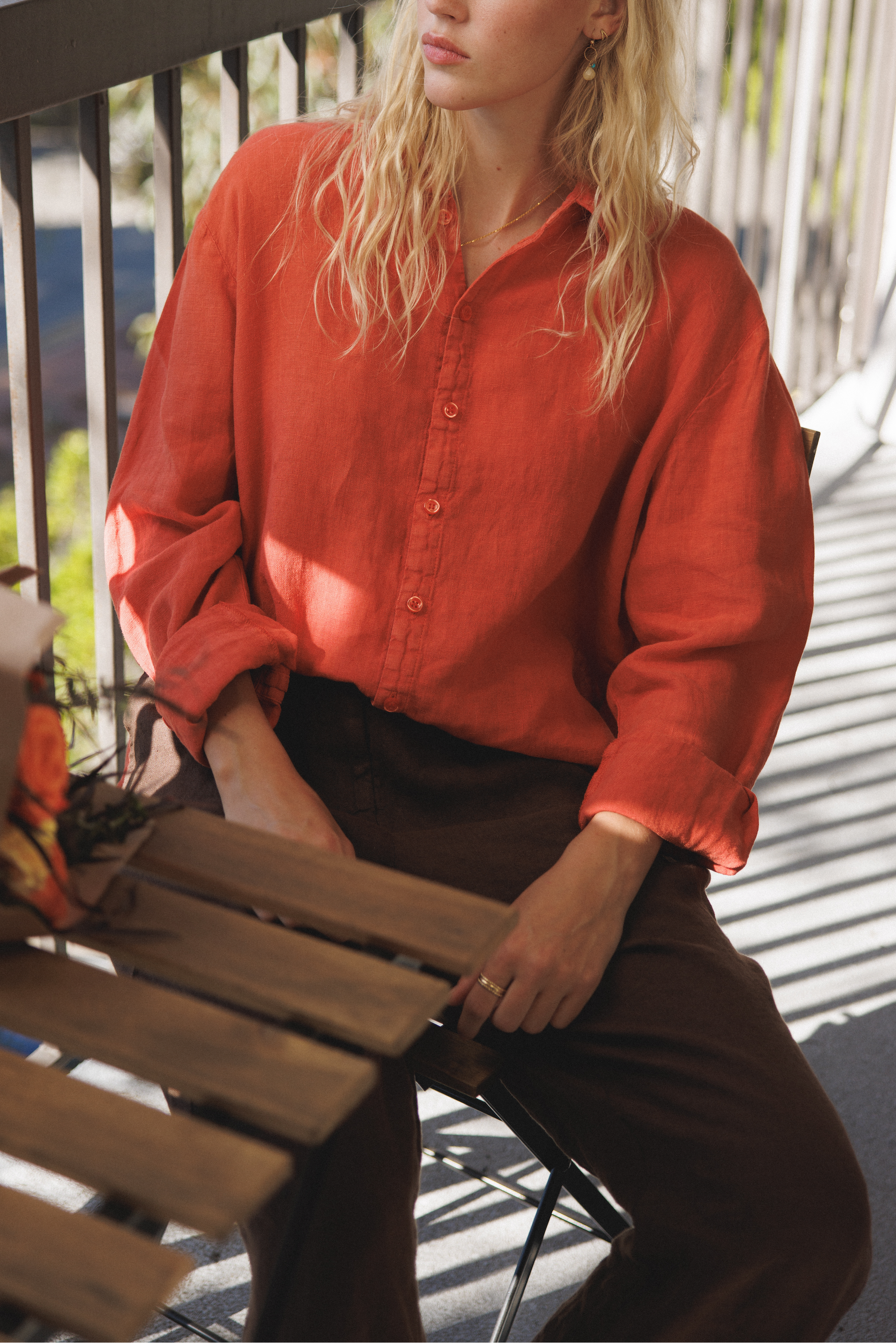 Female model, sitting on a balcony, wearing a cayenne / orange colored sustainable oversized 100% linen button down long sleeve paired with dark brown linen pants - VIR VITA
#color_cayenne