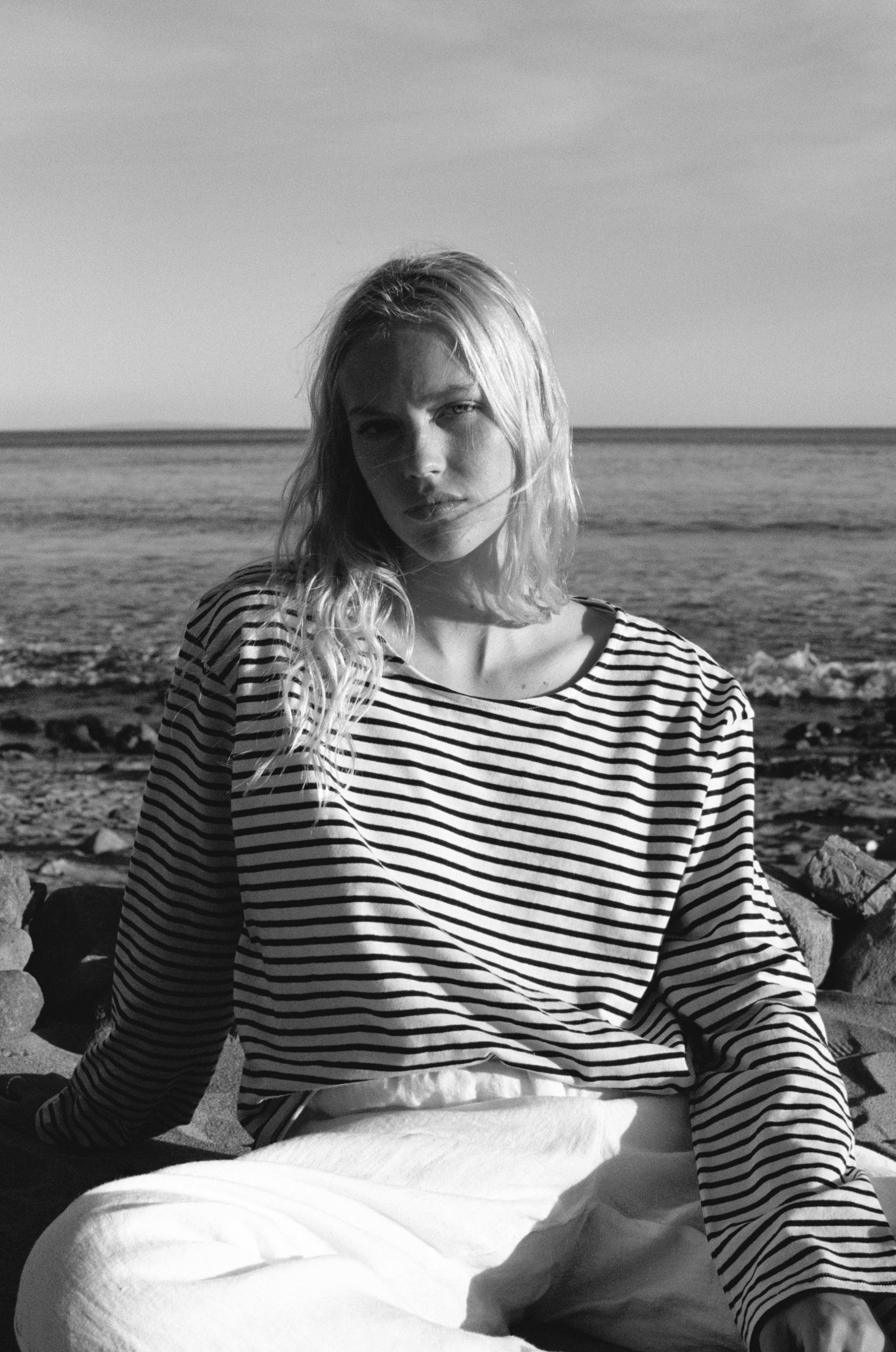 Female model, sitting on the rocks in front of the ocean, wearing a sustainable long sleeve striped navy blue and white top featuring a flattering neckline and oversized fit  - VIR VITA
#color_classic navy stripe