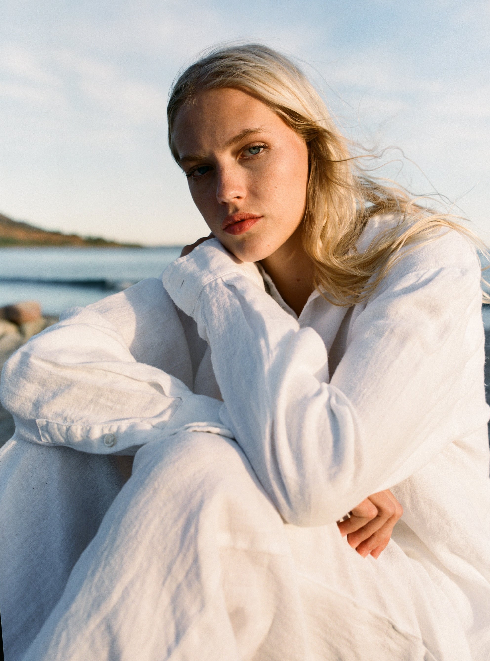 Female model, sitting in front of the ocean, wearing a white sustainable oversized linen button down long sleeve - VIR VITA
#color_white