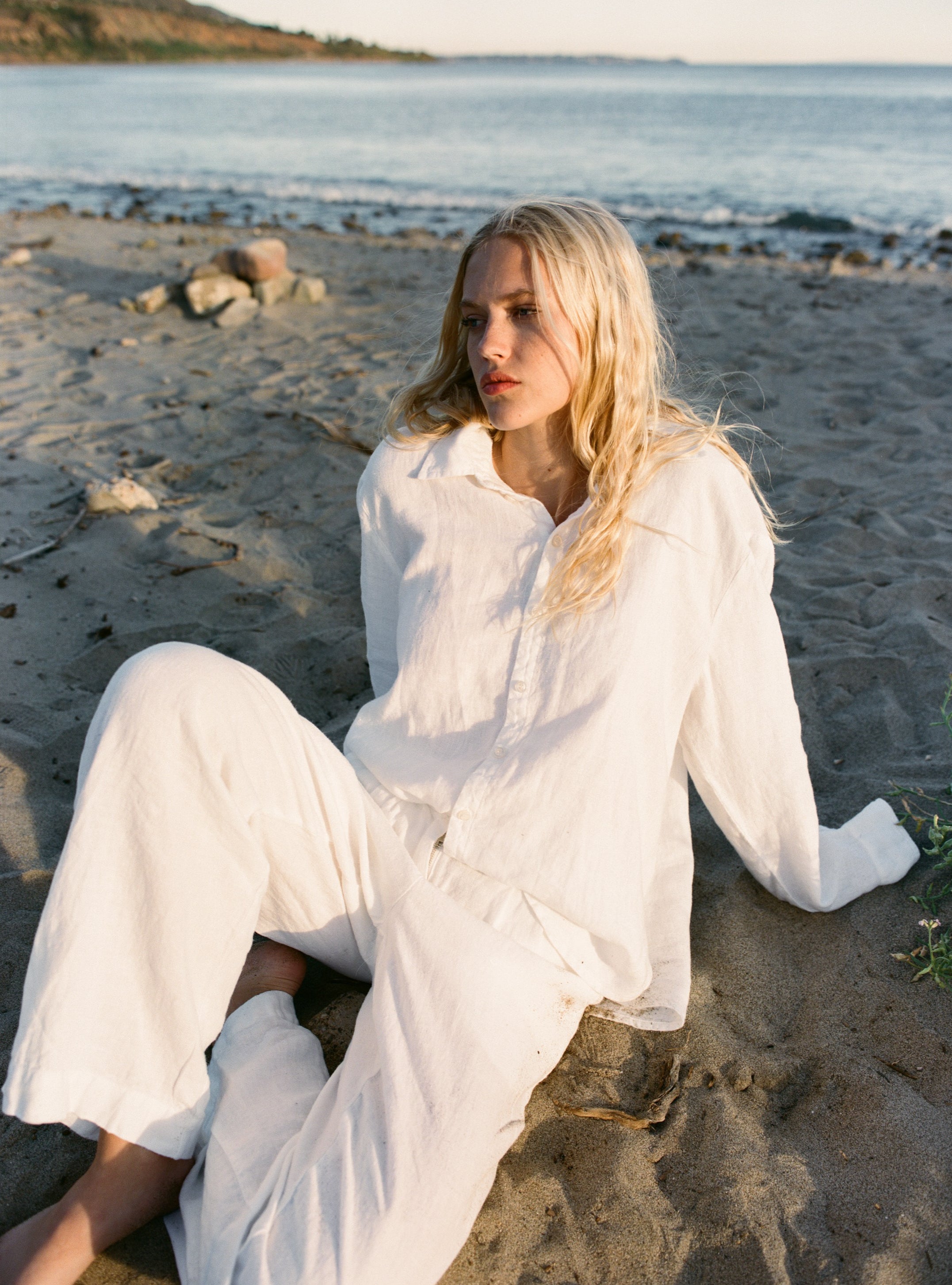 Female model, sitting on the sand in front of the ocean, wearing a white sustainable oversized linen button down long sleeve - VIR VITA
#color_white