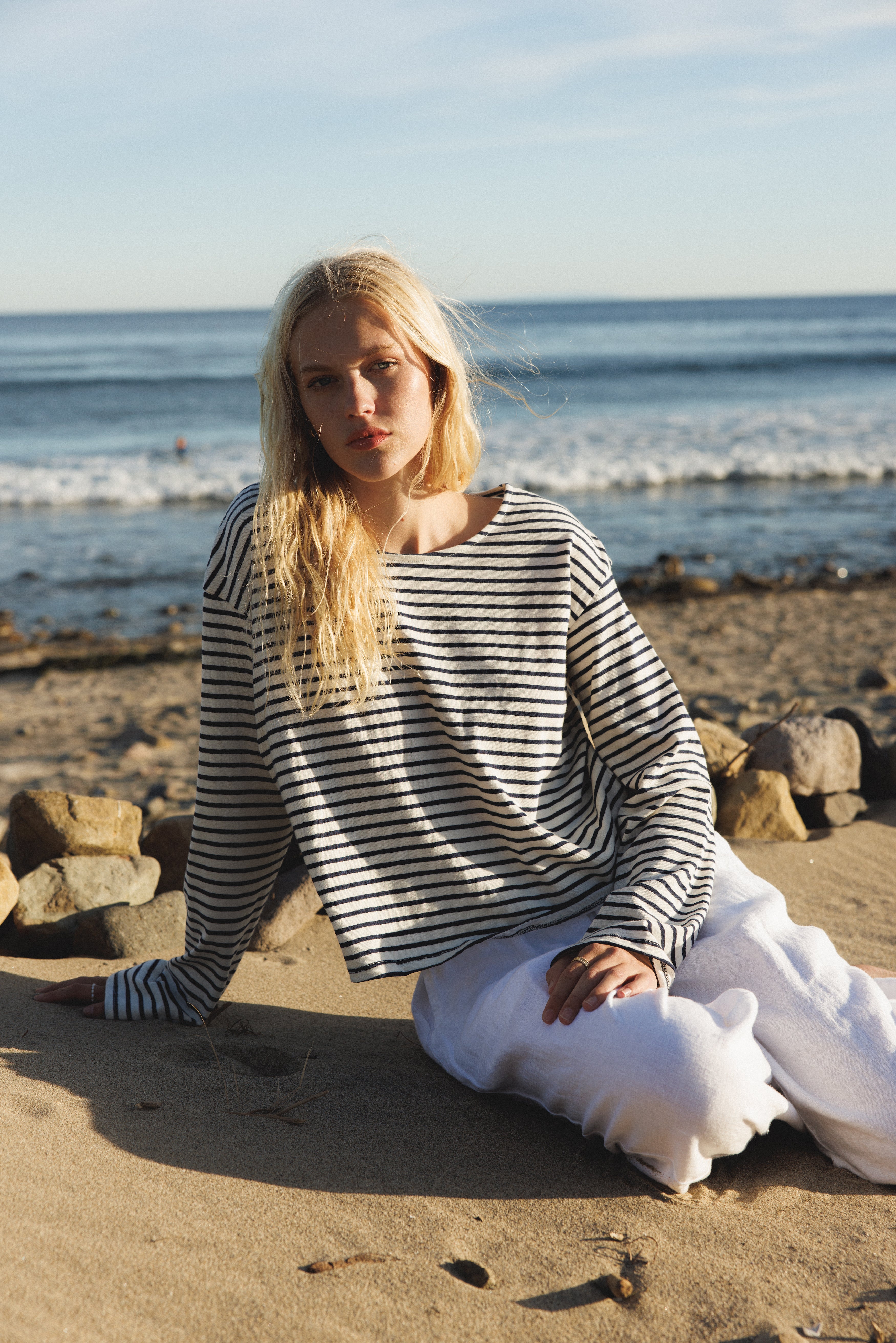 Female model, sitting on the sand in front of the ocean, wearing a sustainable long sleeve striped navy top - VIR VITA