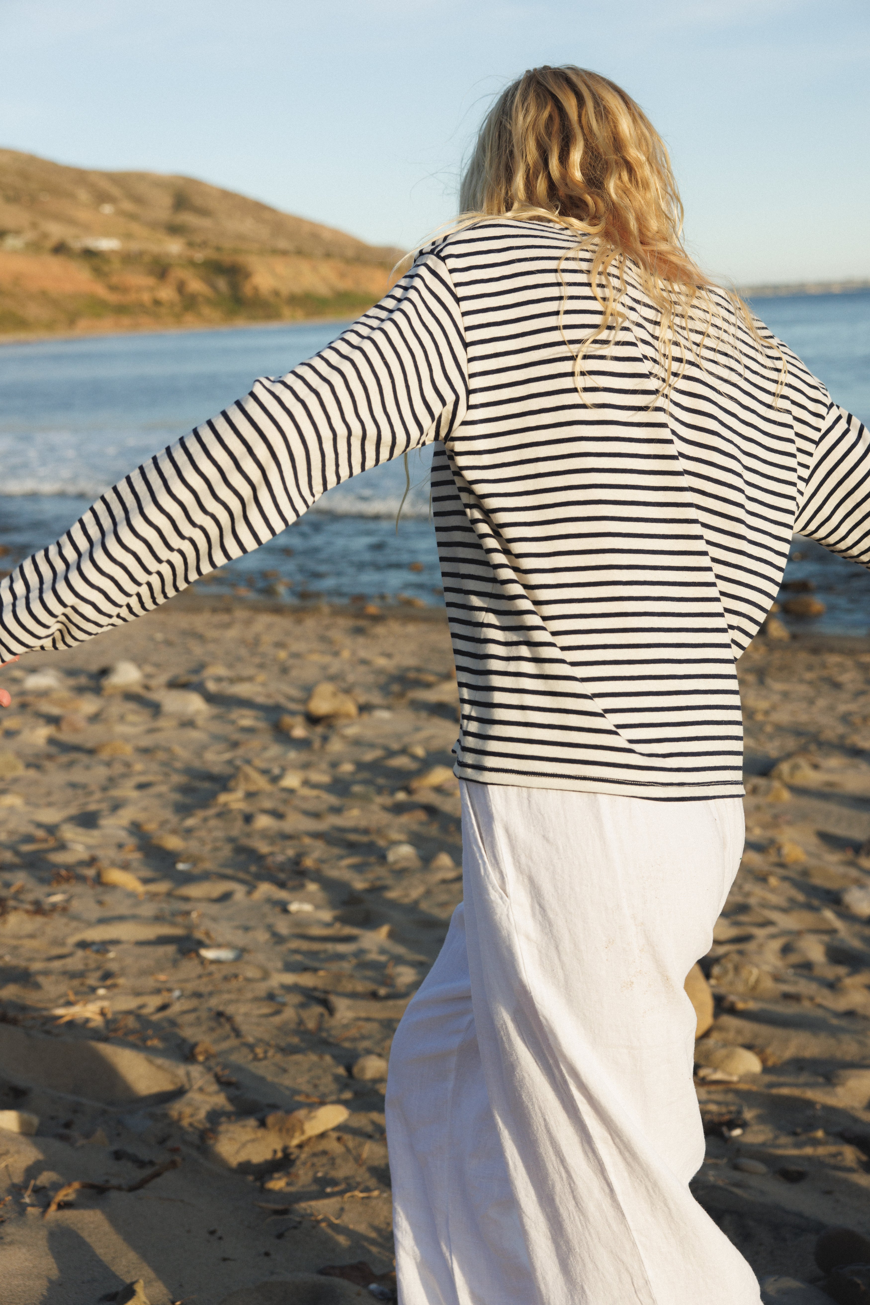 Female model, walking on the sand in front of the ocean, wearing a sustainable long sleeve striped navy top - VIR VITA