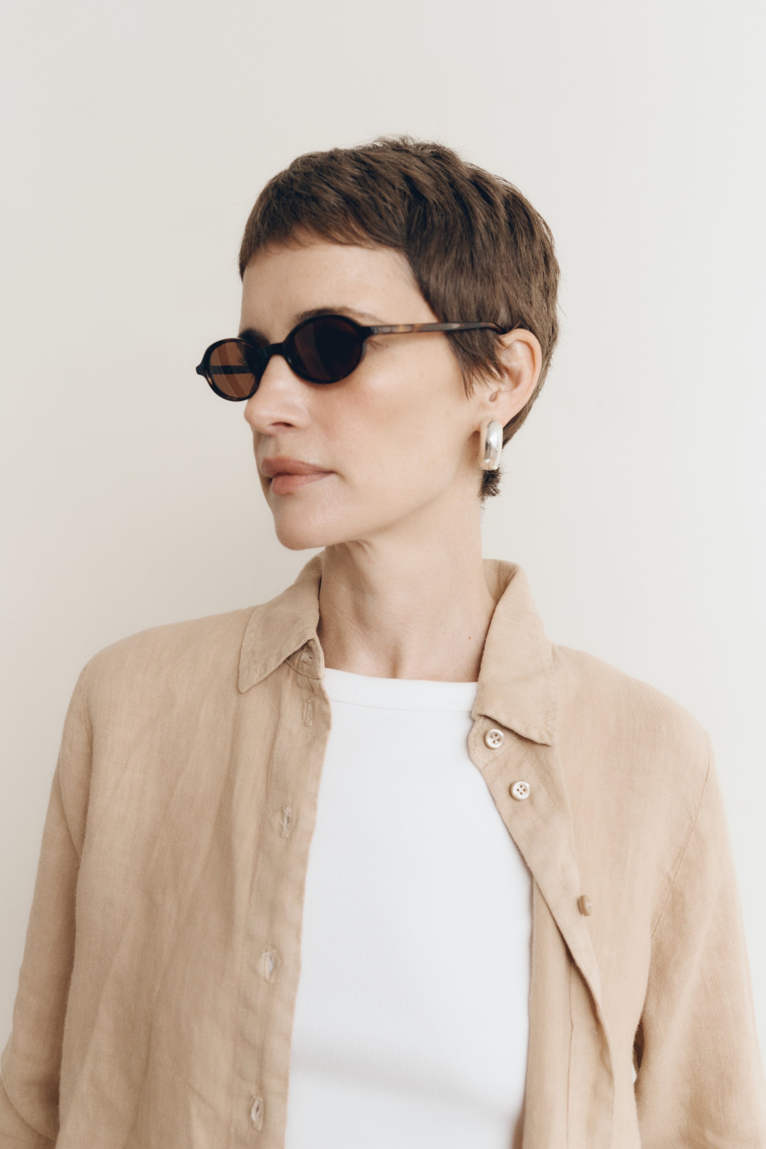 Posed female model wearing a sustainable 100% linen classic button up collared shirt in toffee beige paired a high neck white tank top, and black sunglasses - VIR VITA
#color_toffee