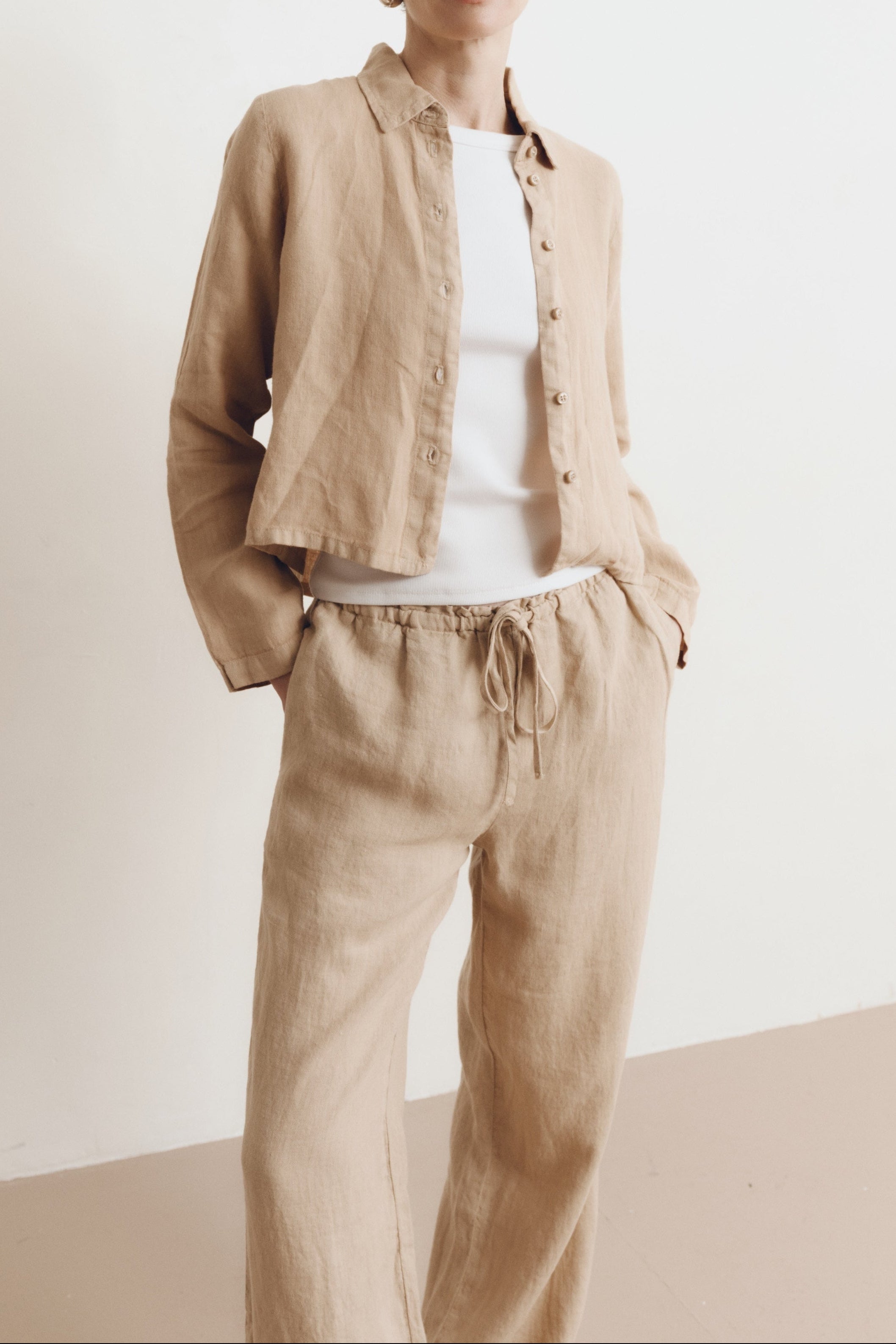 Posed female model wearing a sustainable 100% linen classic button up collared shirt in toffee beige paired with matching drawstring linen pants, a high neck white tank top, and black sunglasses - VIR VITA
#color_toffee