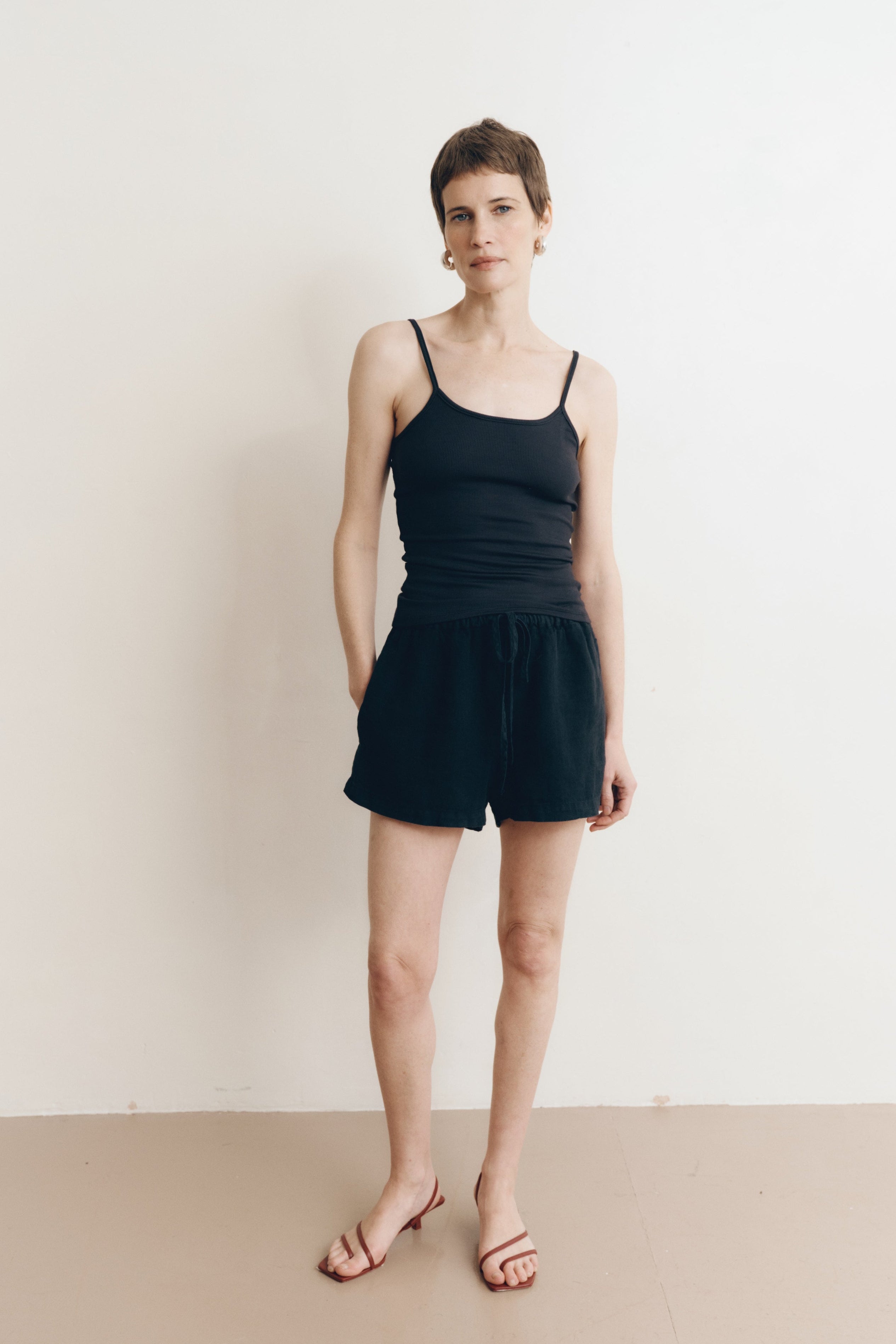 Posed female model wearing sustainable 100% linen drawstring shorts in black paired with a modal cami tank top in black, and brown heels - VIR VITA
#color_black