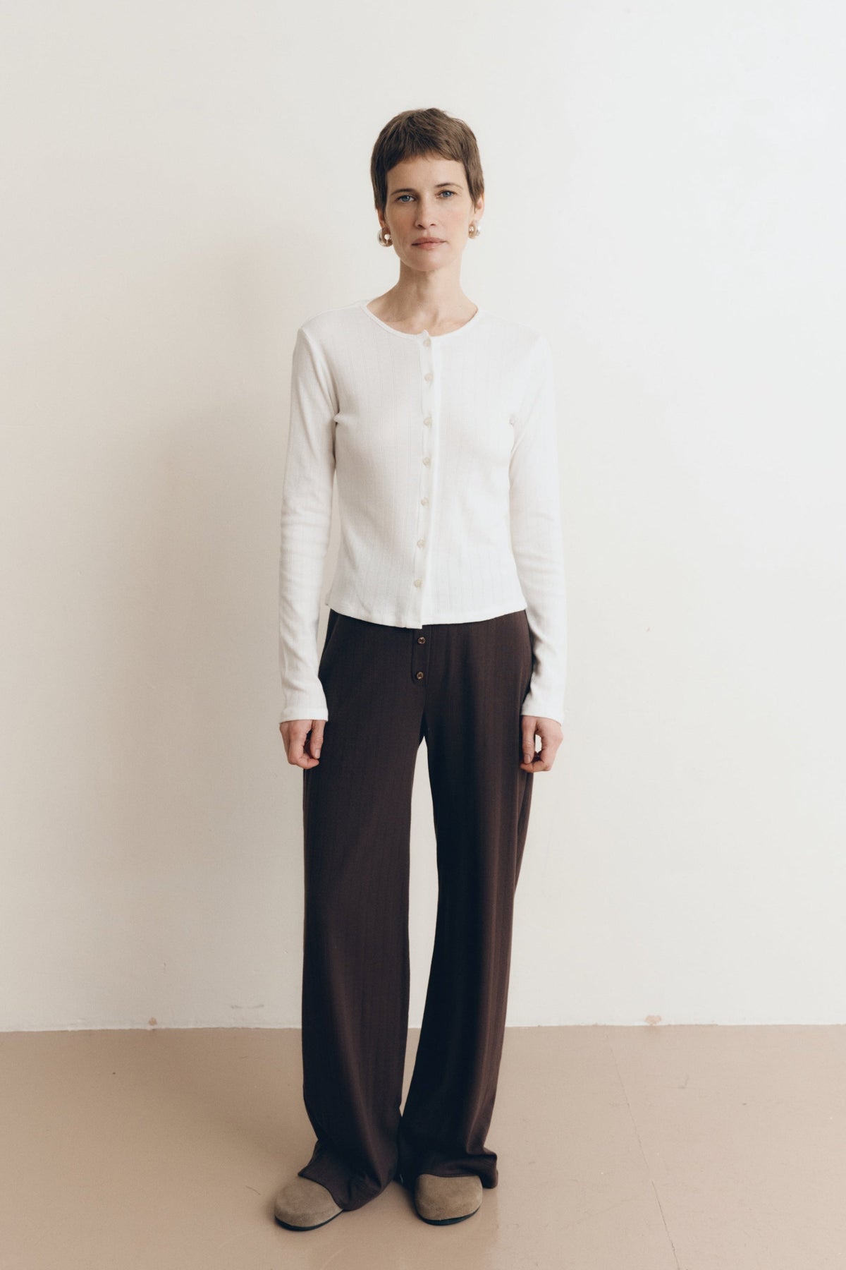 Posed female model wearing a button up sustainable 100% cotton pointelle knit cardigan in white paired with pointelle lounge pants in dark brown and brown suede shoes- VIR VITA
#color_coconut