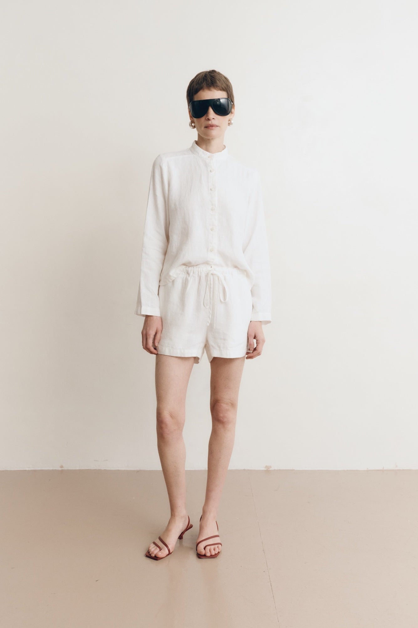 Posed female model wearing sustainable 100% linen drawstring shorts in white paired with a linen button up oversized shirt in white, and red sandals - VIR VITA
#color_white