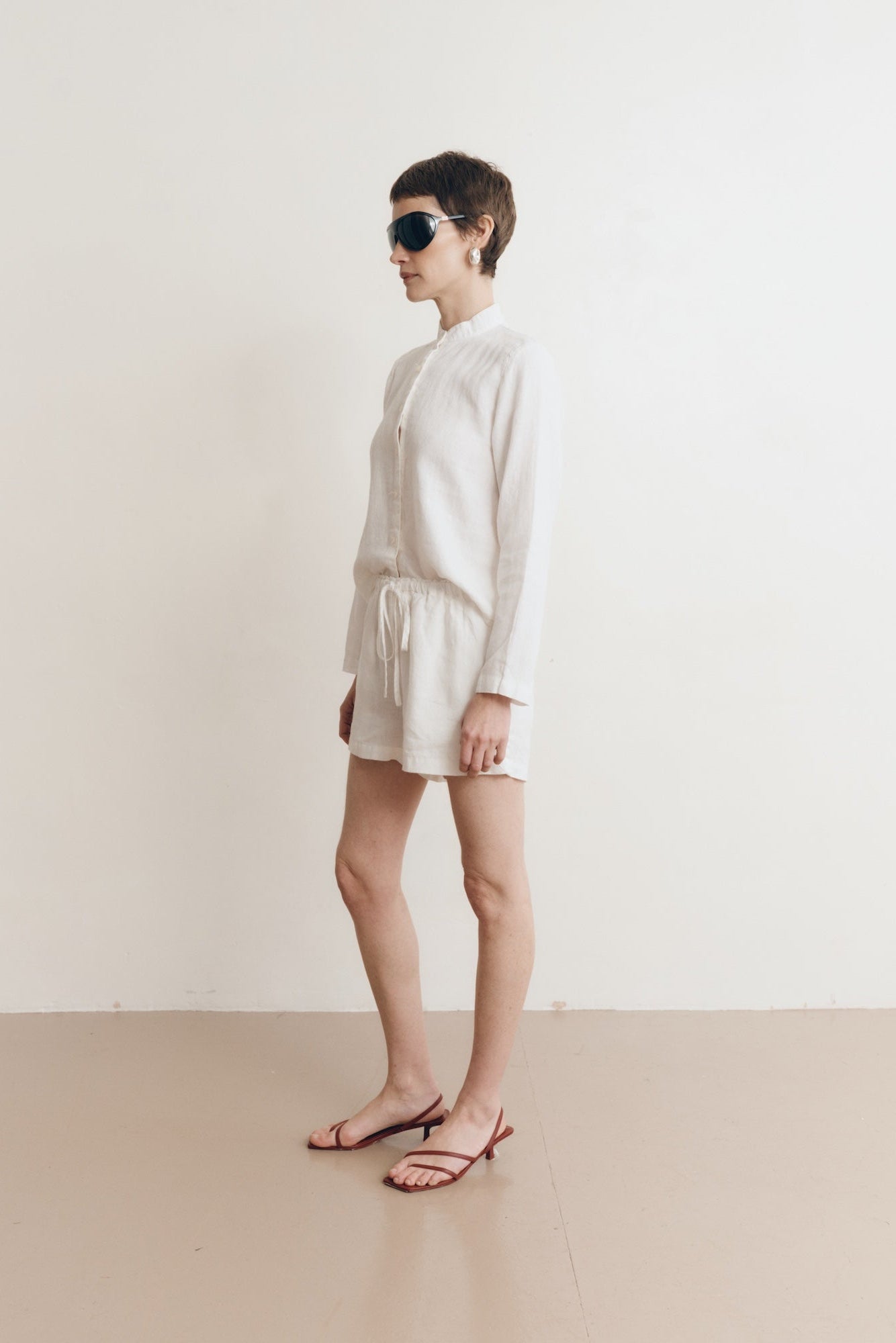 Posed female model wearing sustainable 100% linen drawstring shorts in white paired with a linen button up oversized shirt in white, and red sandals - VIR VITA
#color_white