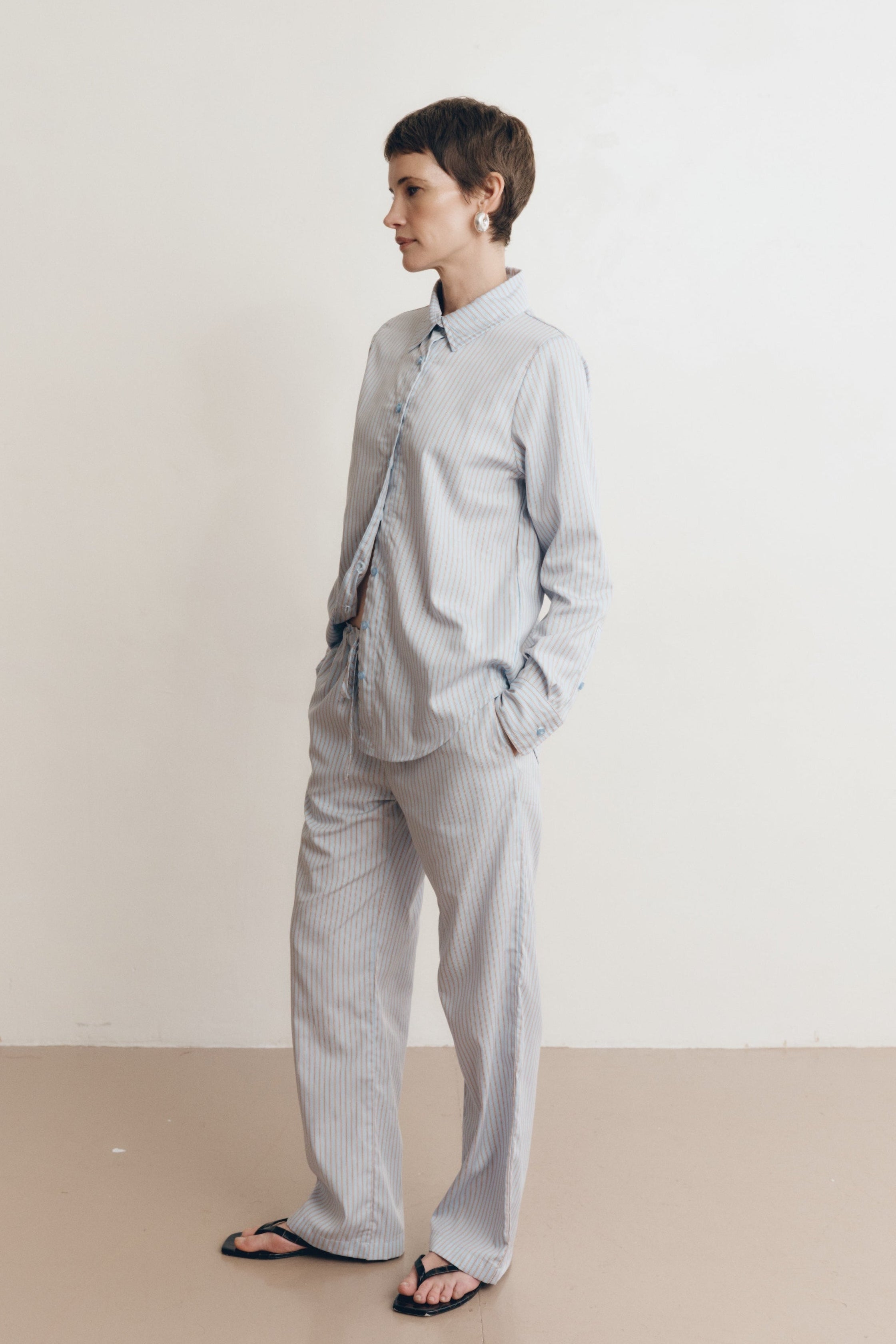 Posed female model wearing a sustainable 100% cotton striped button up collared shirt in light blue paired with matching striped drawstring pants, and black sandals - VIR VITA
#color_seaside stripe