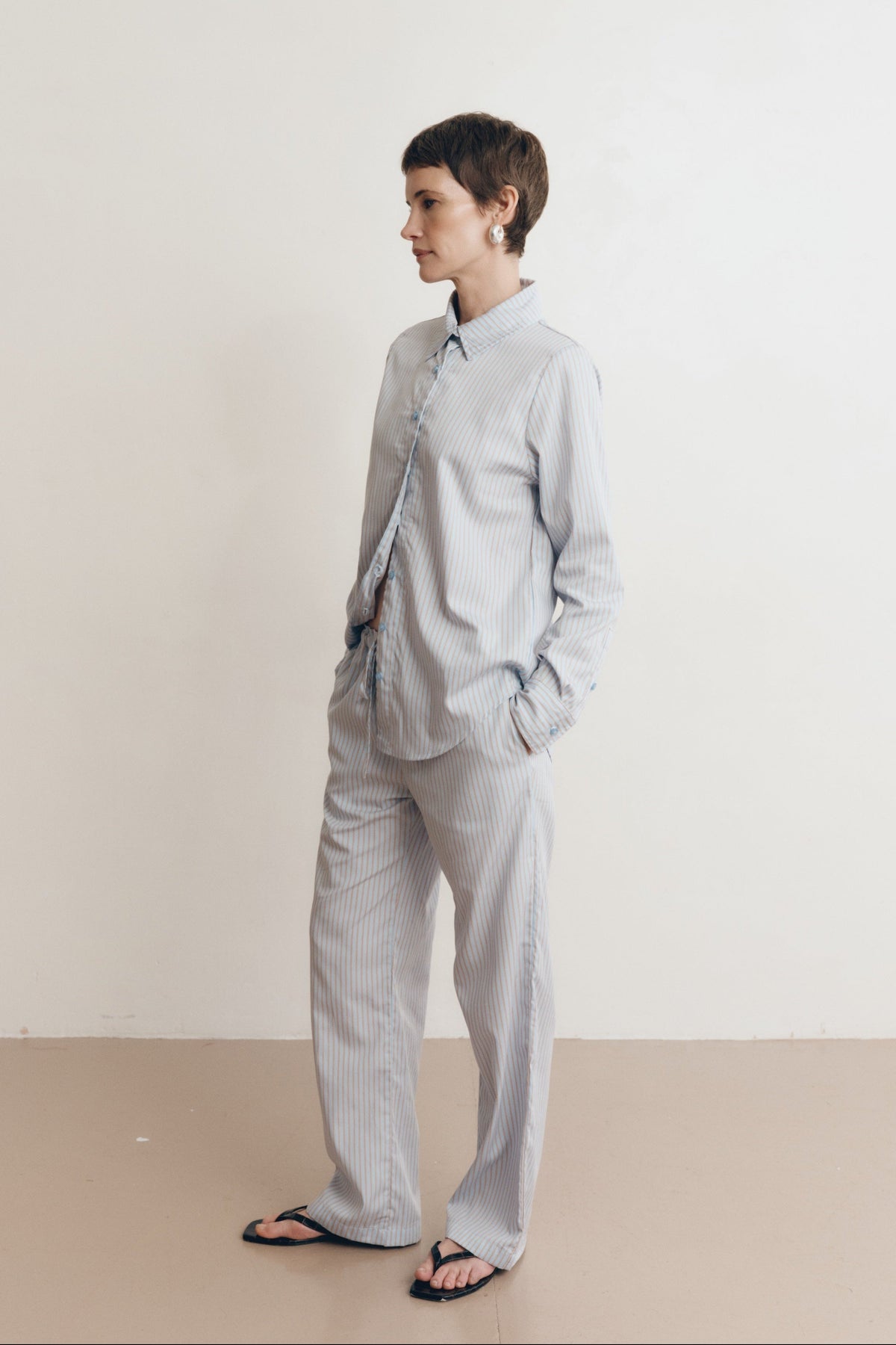 Posed female model wearing sustainable 100% cotton relaxed striped drawstring pants in light blue paired with a matching button up collared shirt in light blue, and black sandals - VIR VITA
#color_seaside stripe