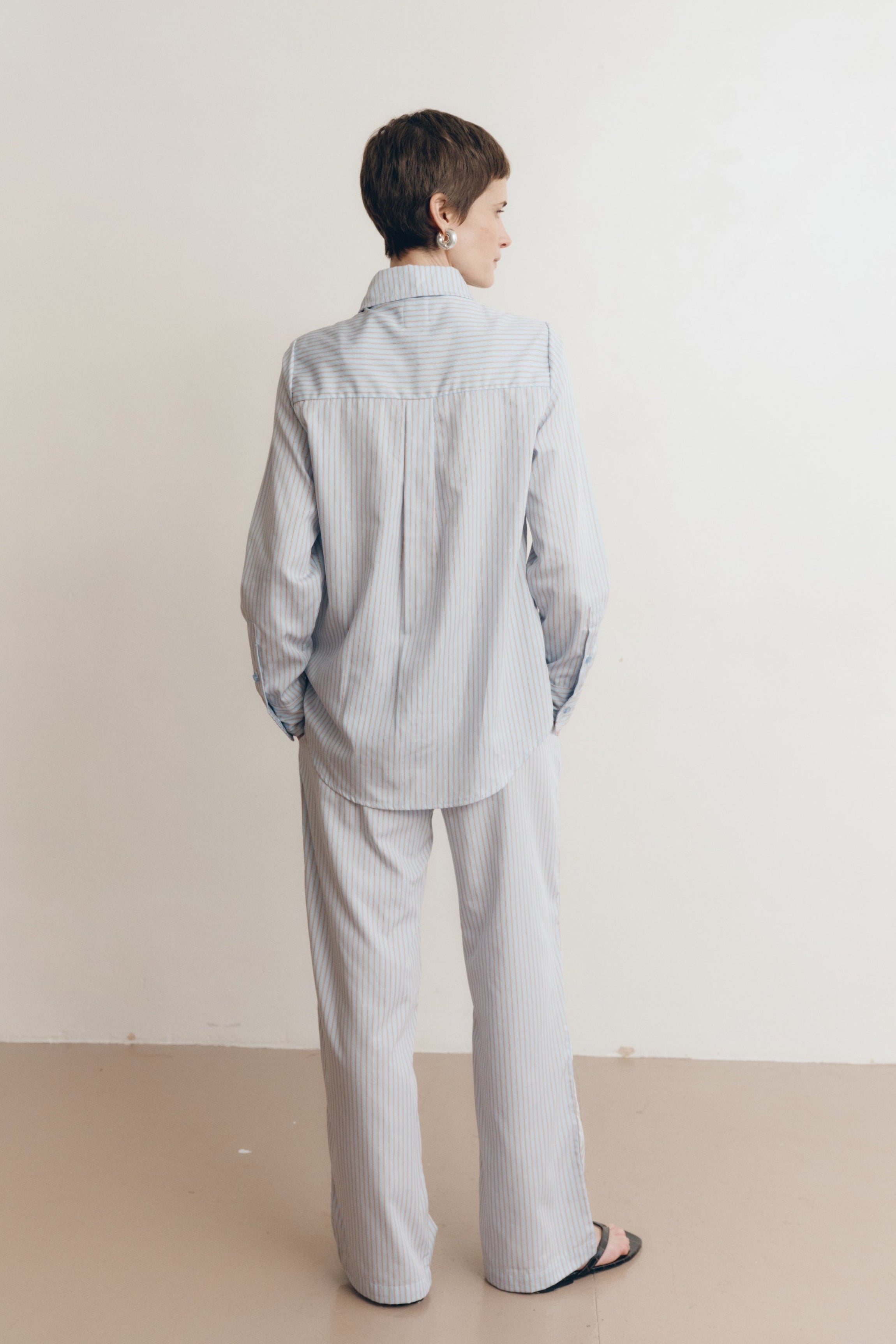 Posed female model wearing a sustainable 100% cotton striped button up collared shirt in light blue paired with matching striped drawstring pants, and black sandals - VIR VITA
#color_seaside stripe