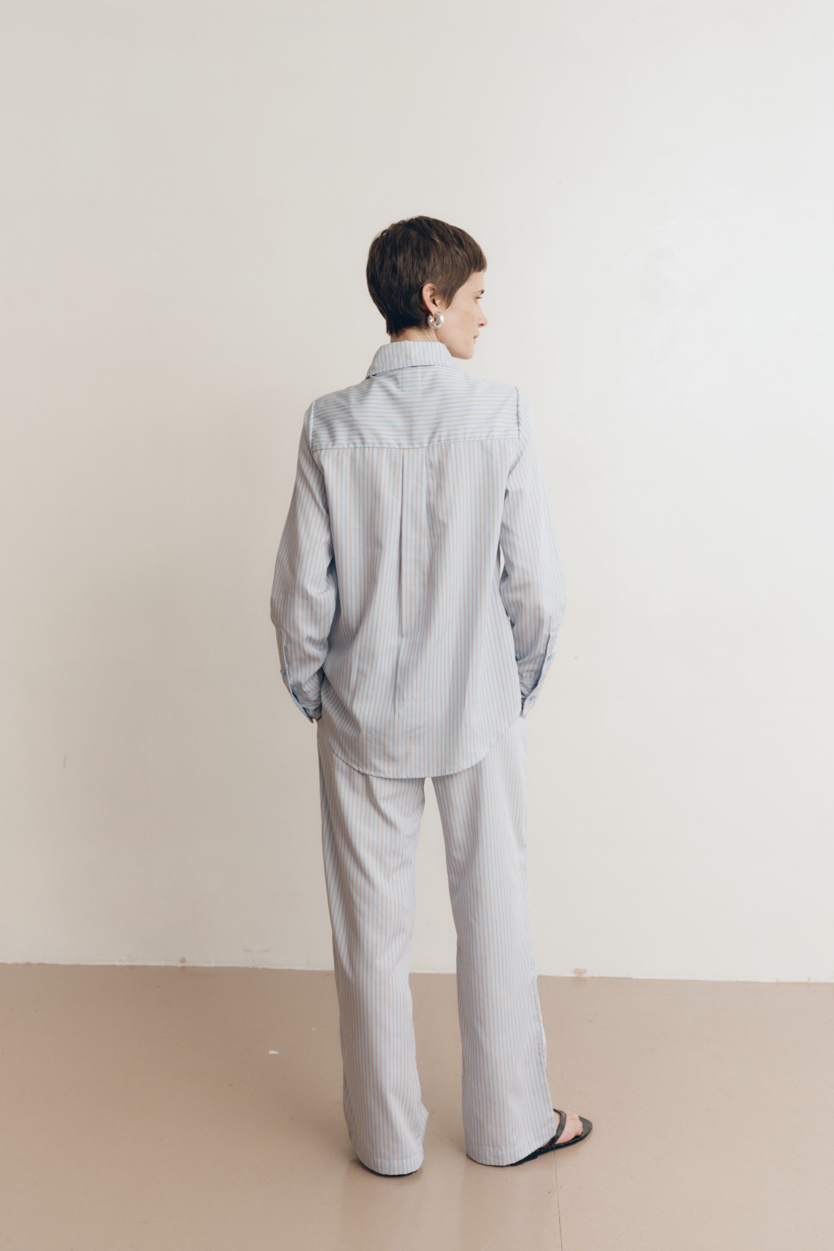 Posed female model wearing sustainable 100% cotton relaxed striped drawstring pants in light blue paired with a matching button up collared shirt in light blue, and black sandals - VIR VITA
#color_seaside stripe