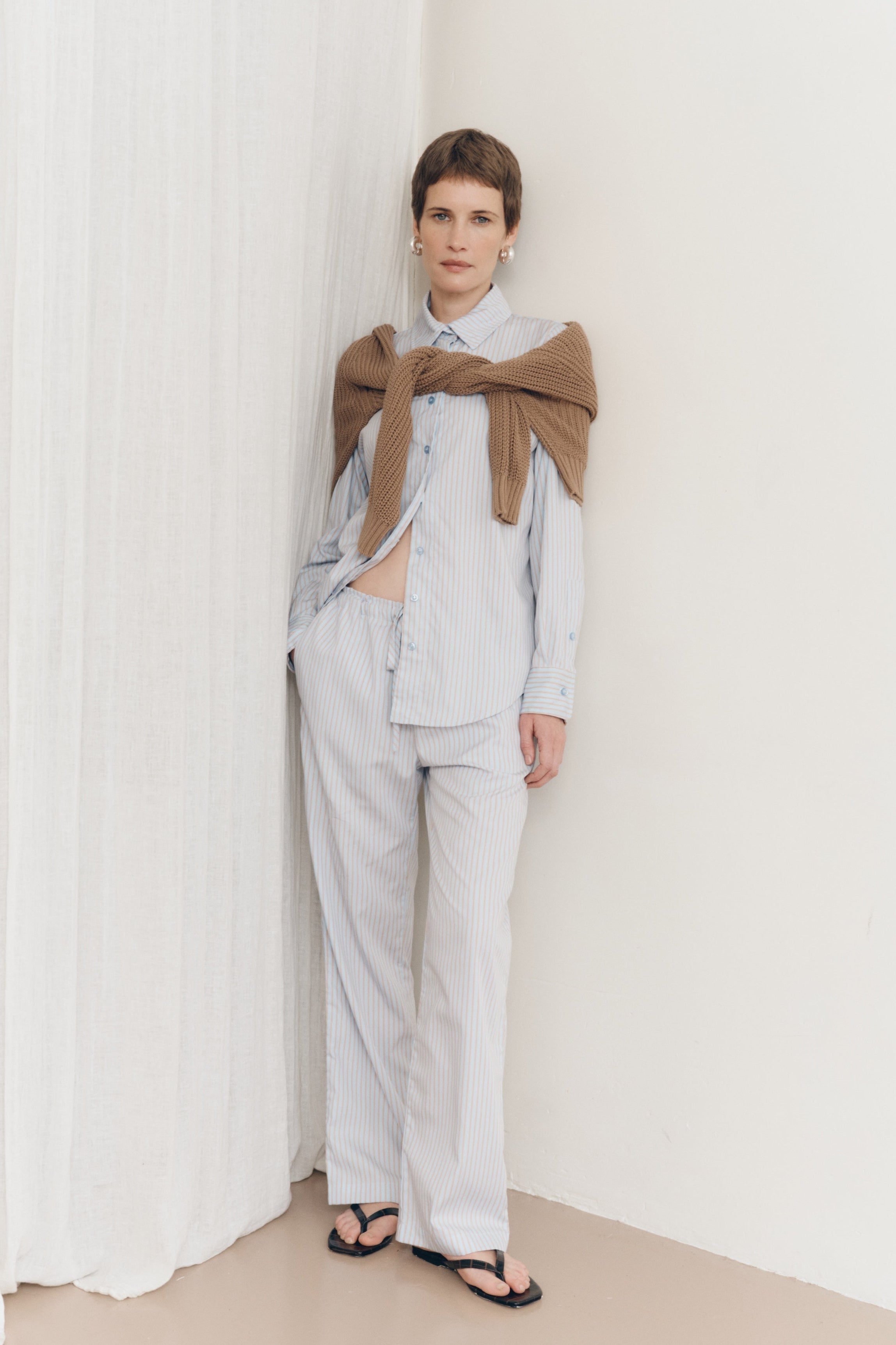 Posed female model wearing a sustainable 100% cotton striped button up collared shirt in light blue paired with matching striped drawstring pants, a light brown cotton knit sweater, and black sandals - VIR VITA
#color_seaside stripe