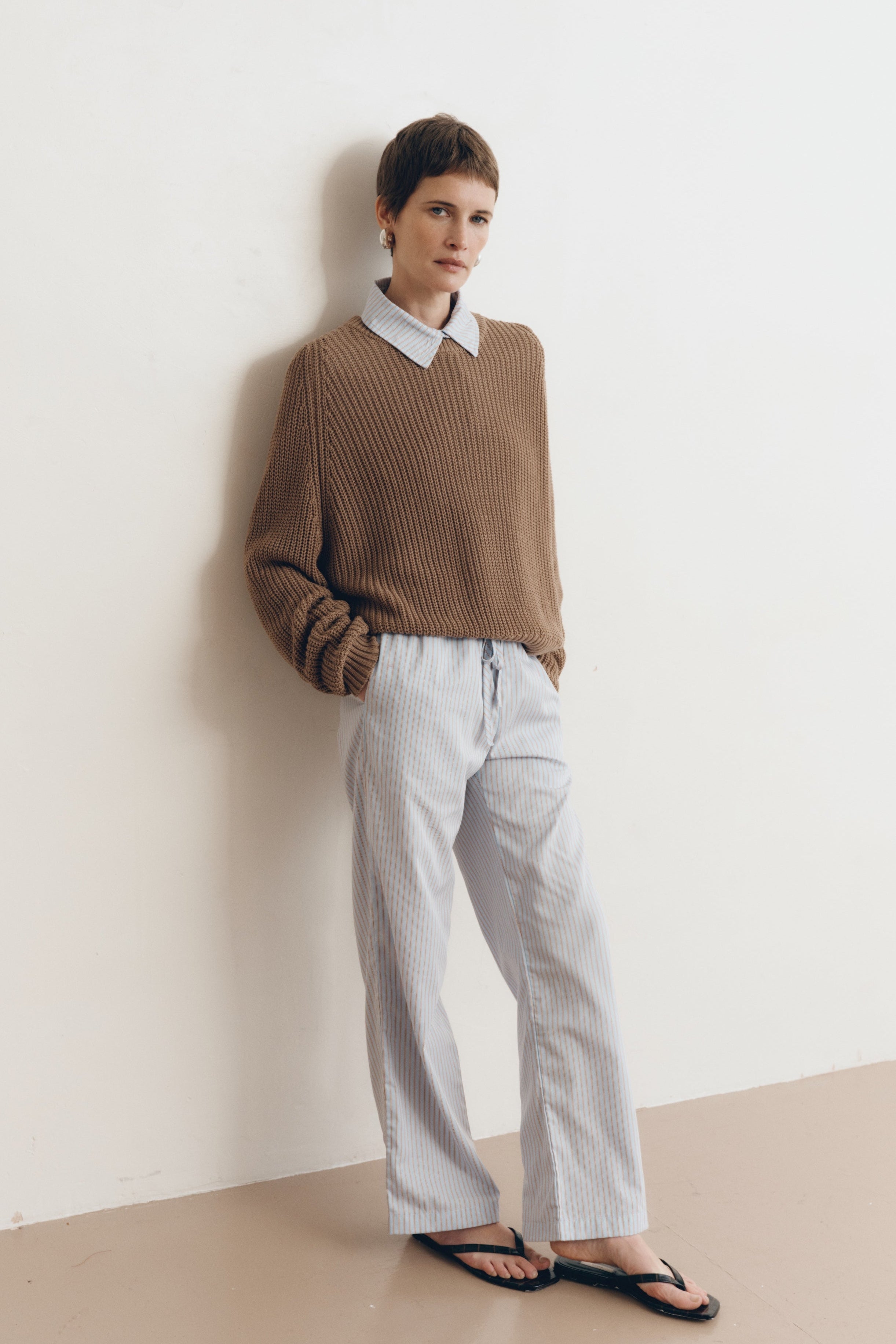 Posed female model wearing a 100% cotton knit sweater with a flattering and subtly cropped silhouette in light brown paired with a striped collared shirt in light blue and matching striped drawstring pants with black sandals - VIR VITA
#color_bark