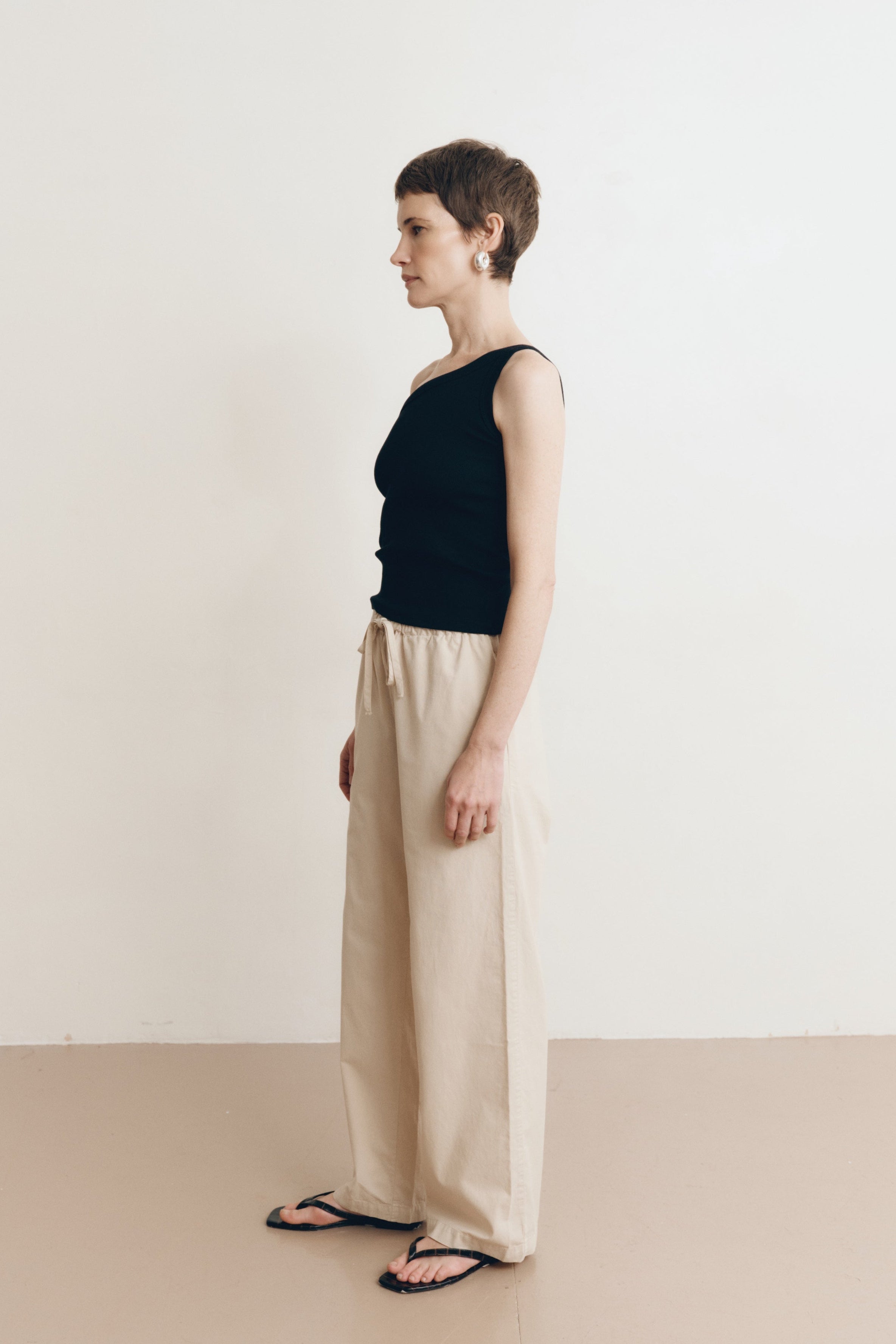 Posed female model wearing 100% cotton poplin drawstring relaxed pants in beige paired with a one shoulder tank top in black and black sandals - VIR VITA
#color_dune