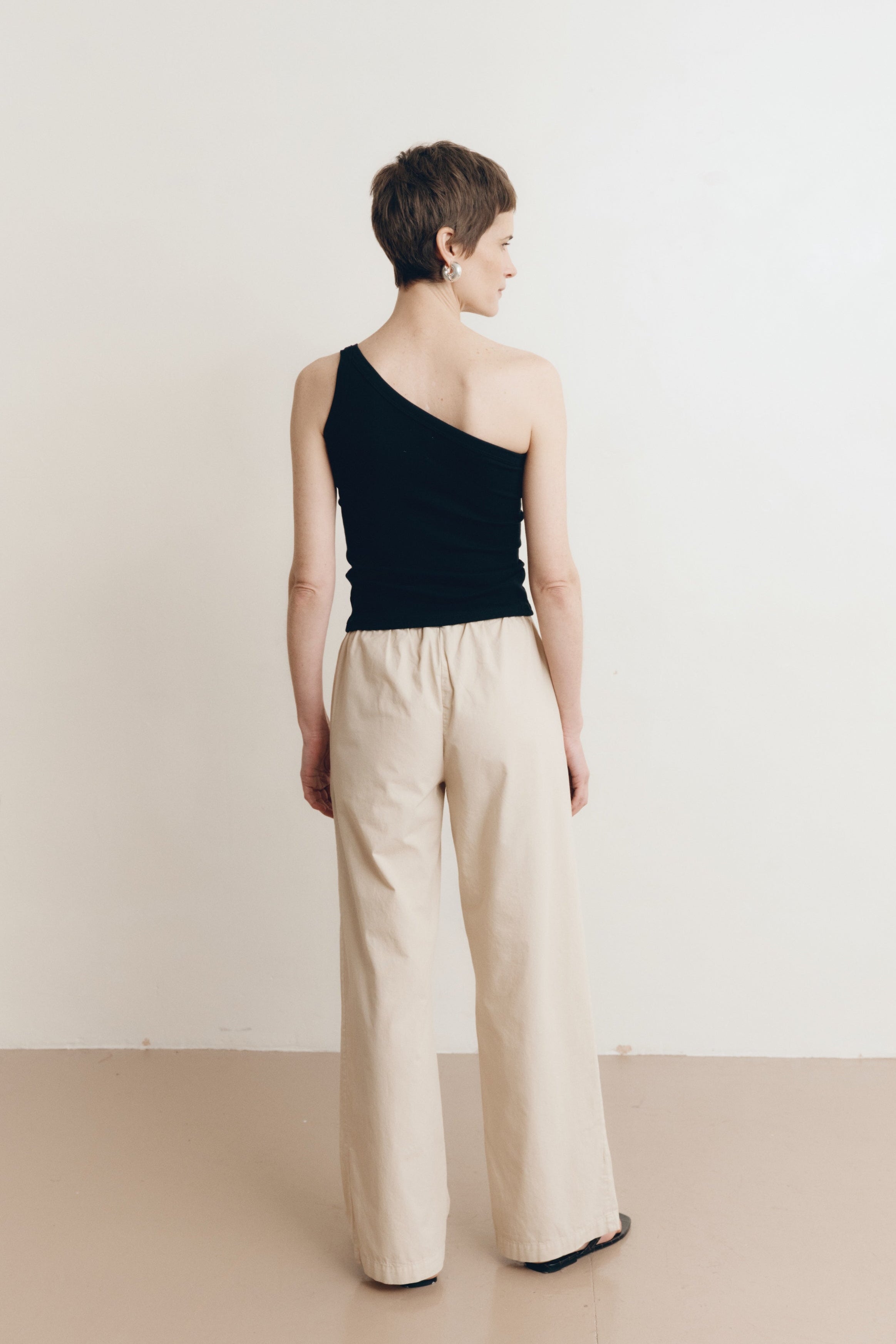 Posed female model wearing 100% cotton poplin drawstring relaxed pants in beige paired with a one shoulder tank top in black and black sandals - VIR VITA
#color_dune