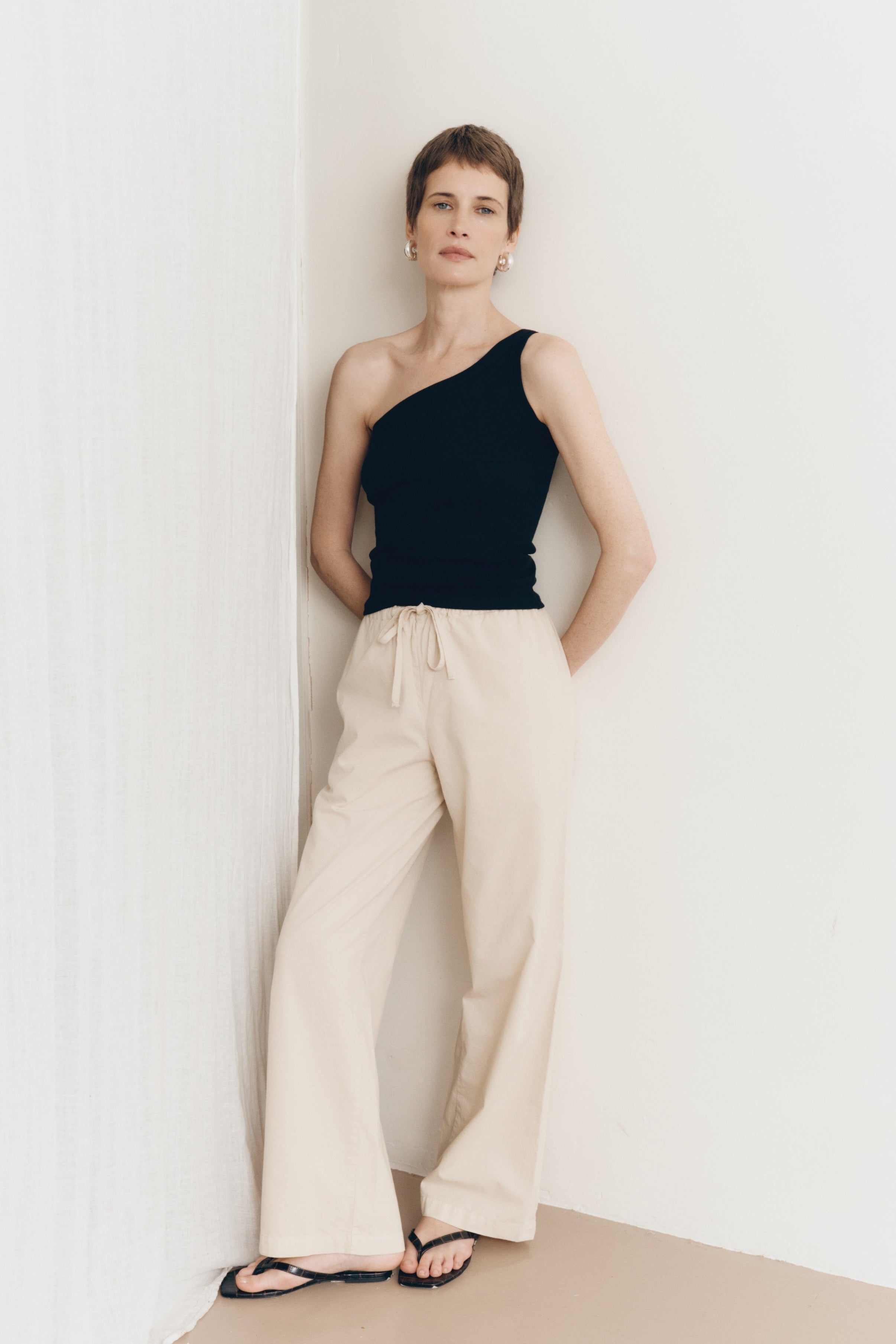 Posed female model wearing 100% cotton poplin drawstring relaxed pants in beige paired with a one shoulder tank top in black and black sandals - VIR VITA
#color_dune
