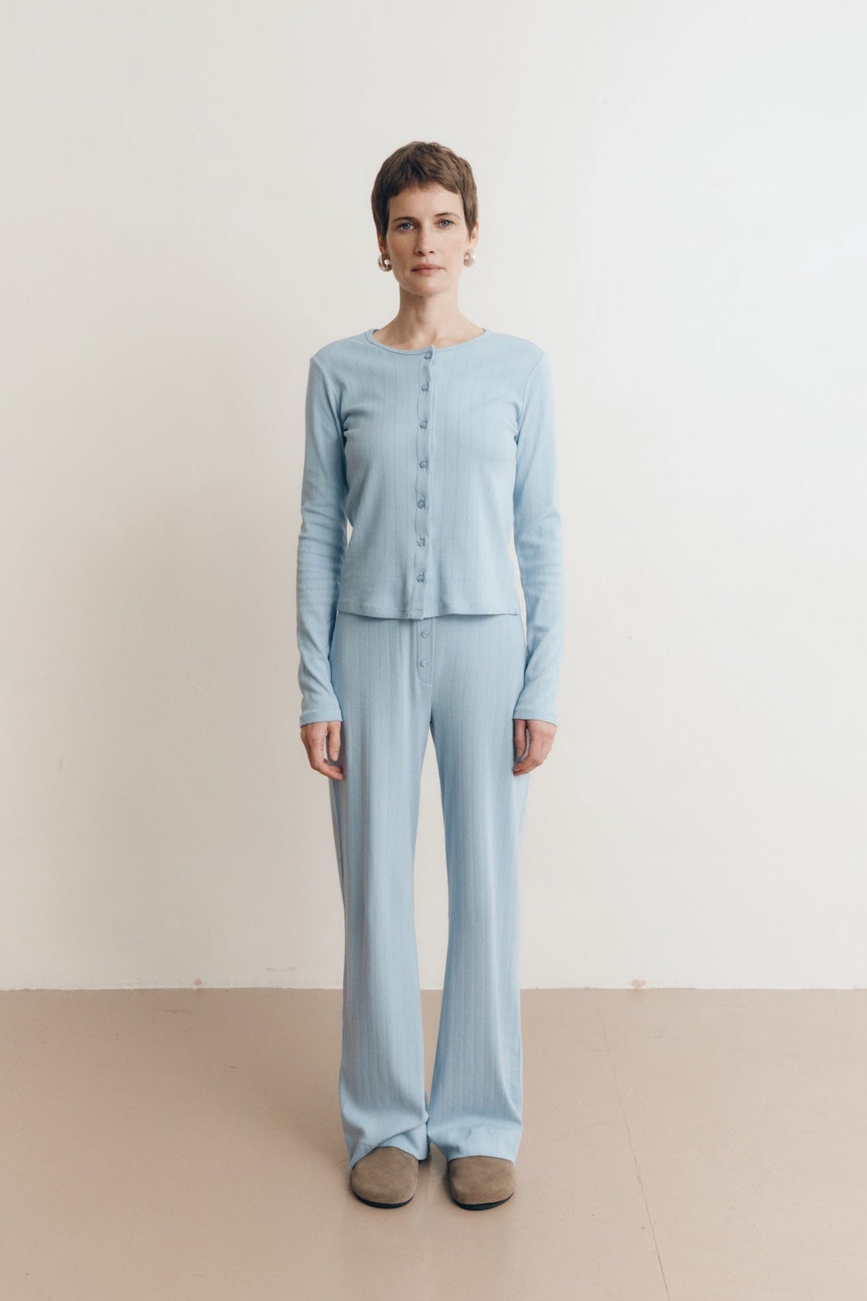 Posed female model wearing a button up sustainable 100% cotton pointelle knit cardigan in light blue paired with a matching pointelle scoop neck tee and pointelle lounge pants with brown suede shoes- VIR VITA
#color_vintage blue