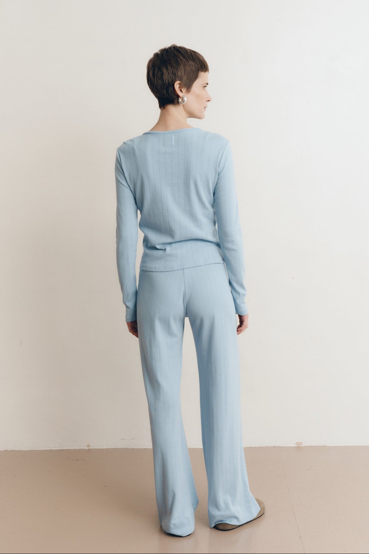 Posed female model wearing a button up sustainable 100% cotton pointelle knit cardigan in light blue paired with matching pointelle lounge pants with brown suede shoes- VIR VITA
#color_vintage blue