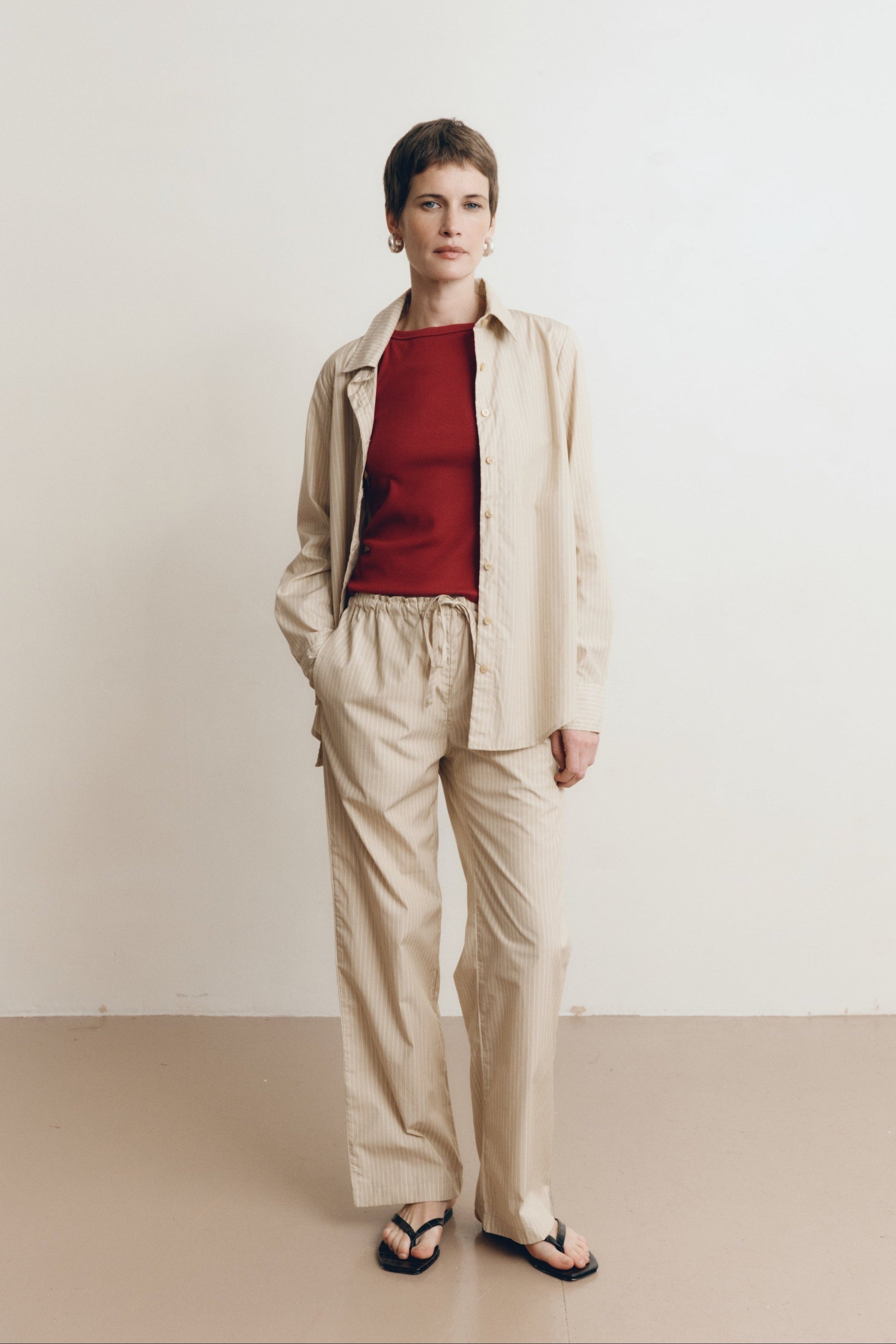 Posed female model wearing a sustainable 100% cotton striped button up collared shirt in beige paired with matching striped drawstring pants, a high neck tank top in dark red, and black sandals - VIR VITA
#color_dune stripe