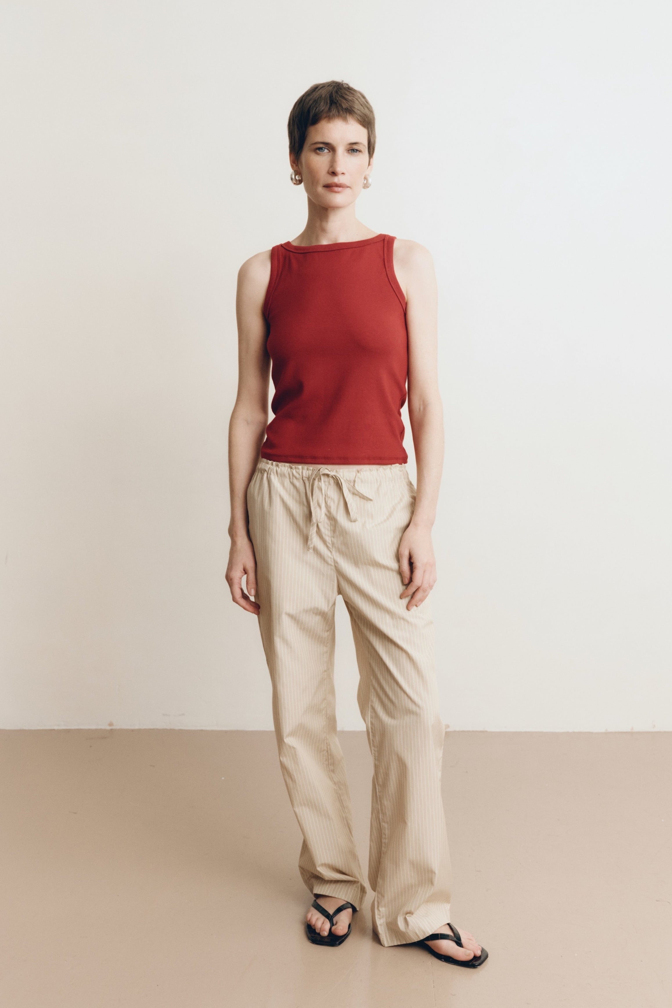 Posed female model wearing sustainable 100% cotton relaxed striped drawstring pants in beige paired with a high neck dark red tank top, and black sandals - VIR VITA
#color_dune stripe