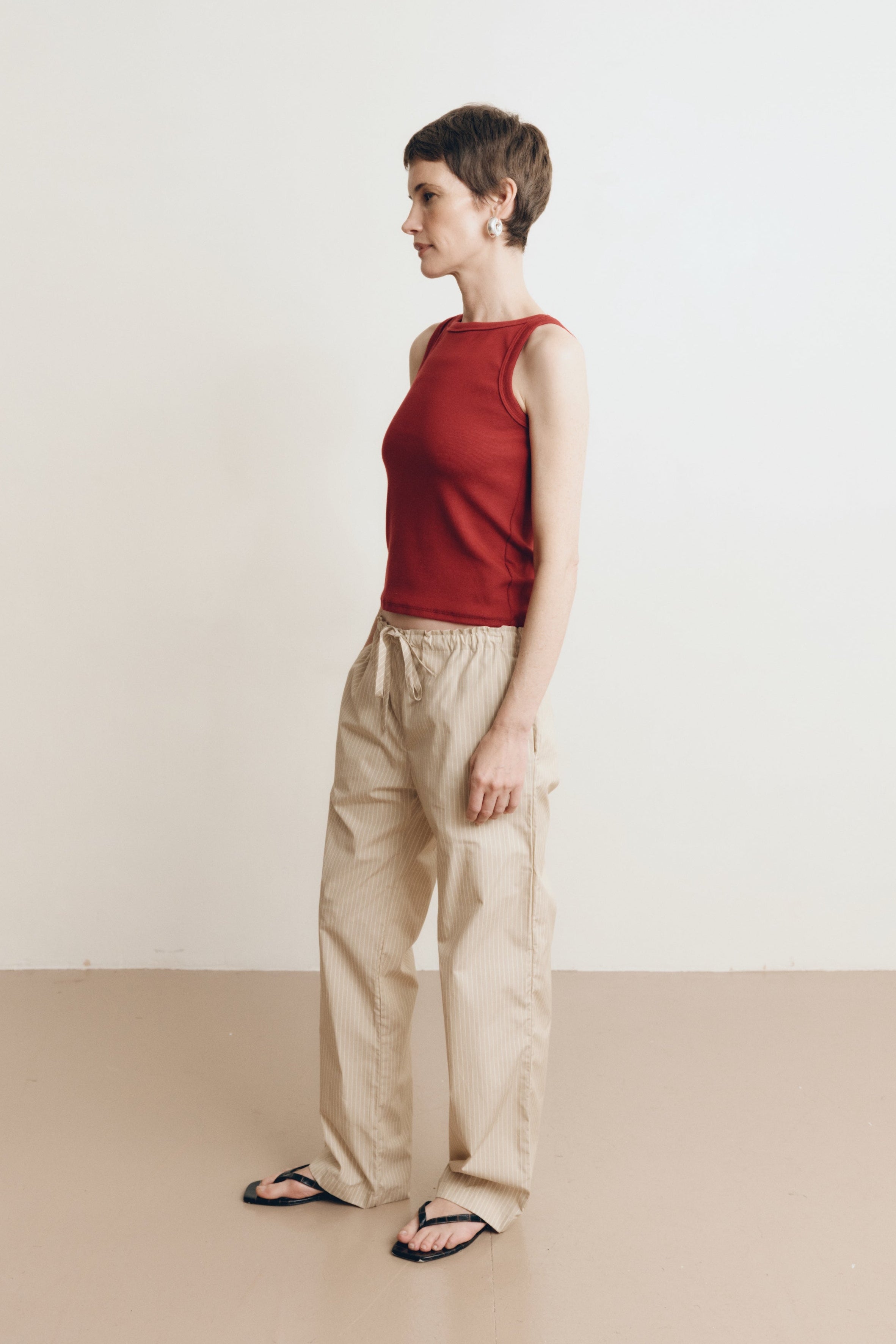 Posed female model wearing sustainable 100% cotton relaxed striped drawstring pants in beige paired with a high neck dark red tank top, and black sandals - VIR VITA
#color_dune stripe