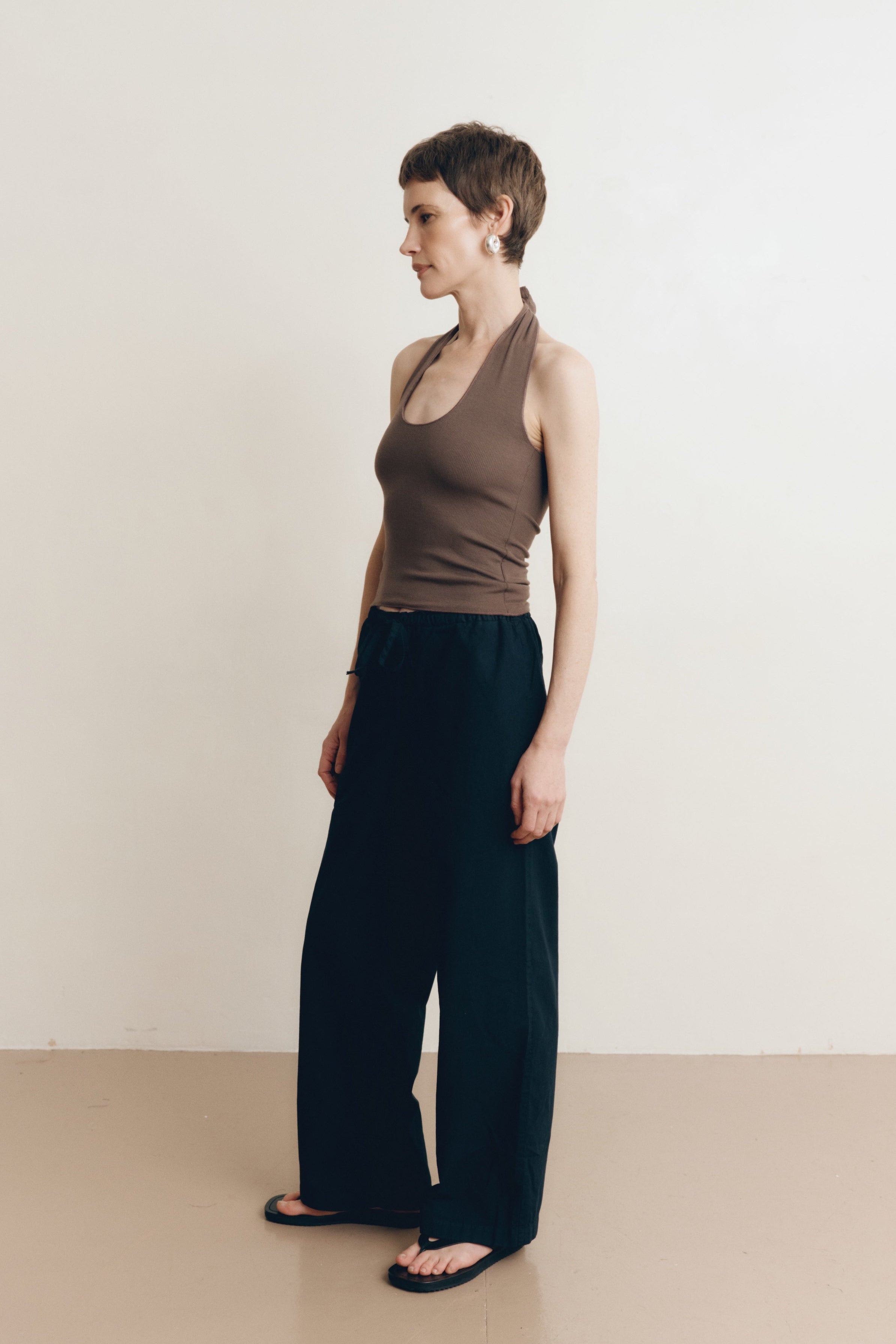 Posed female model wearing 100% cotton poplin drawstring relaxed pants in black paired with a modal dark brown halter top and black sandals - VIR VITA
#color_black