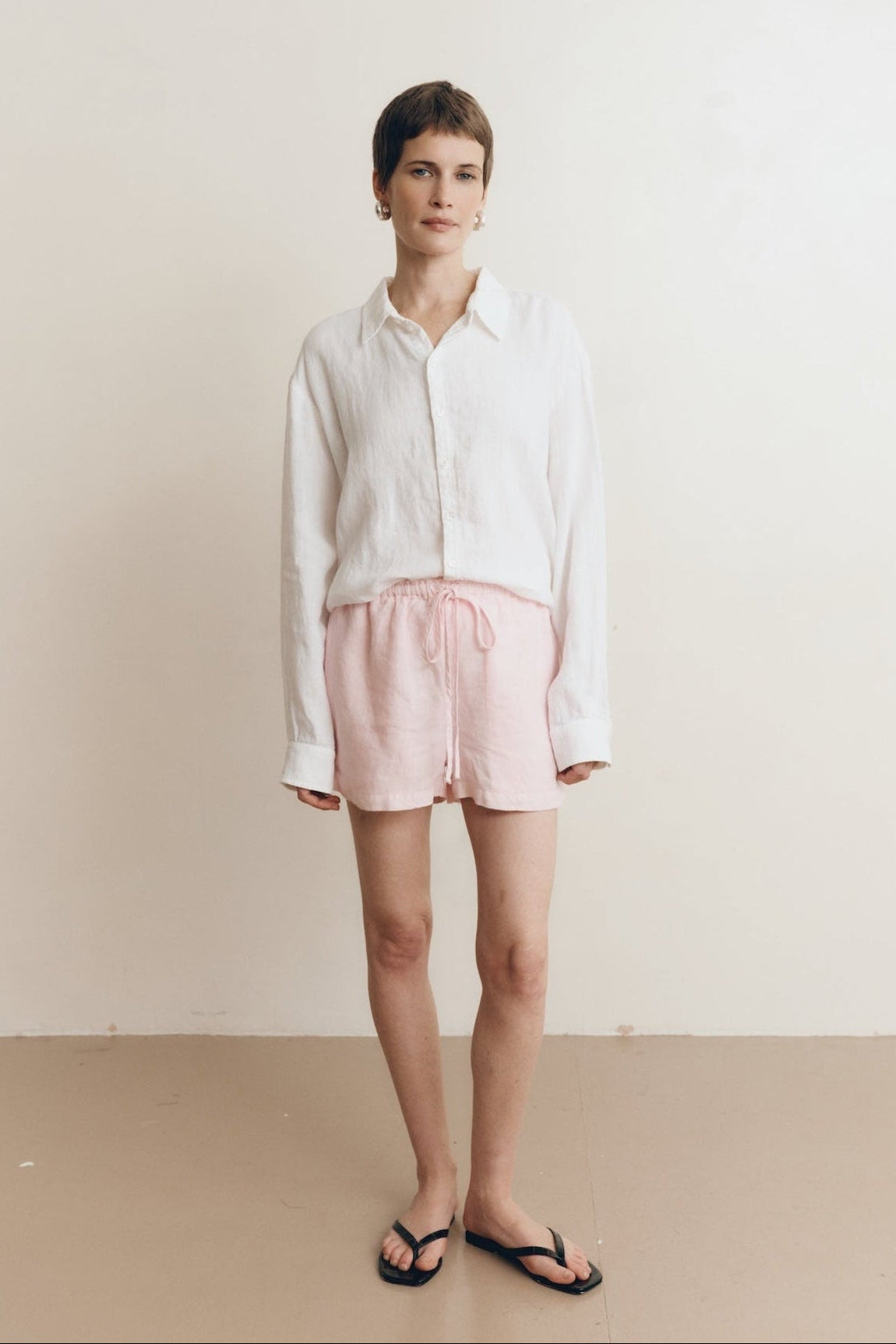 Posed female model wearing sustainable 100% linen drawstring shorts in light pink paired with a linen button up oversized shirt in white, and black sandals - VIR VITA
#color_blossom