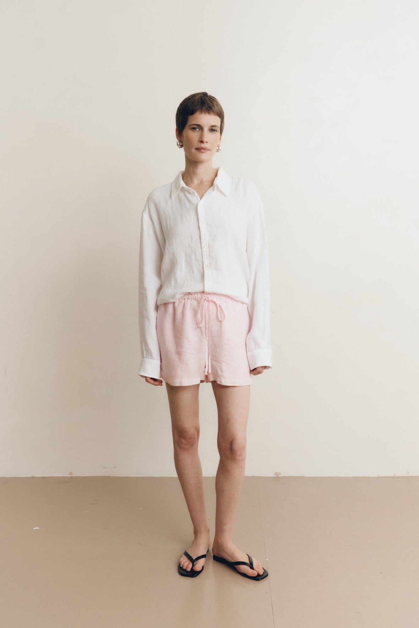Posed female model wearing sustainable 100% linen drawstring shorts in light pink paired with a linen button up oversized shirt in white, and black sandals - VIR VITA
#color_blossom