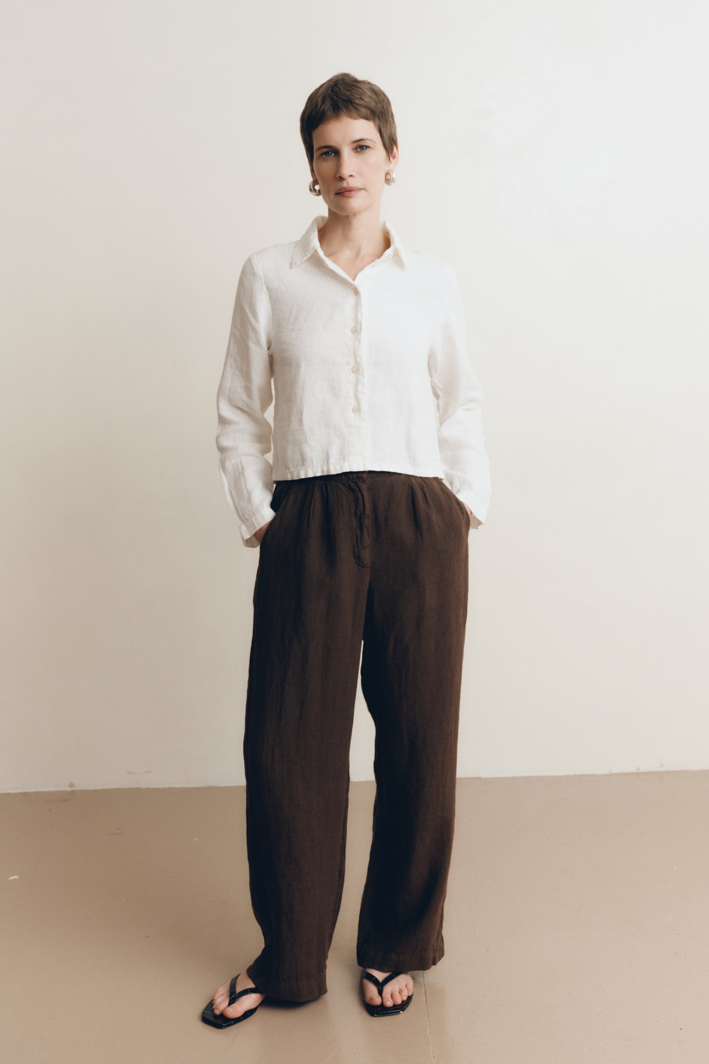 Posed female model wearing a sustainable 100% linen classic button up collared shirt in white paired with dark brown linen trousers and black sandals- VIR VITA
#color_white