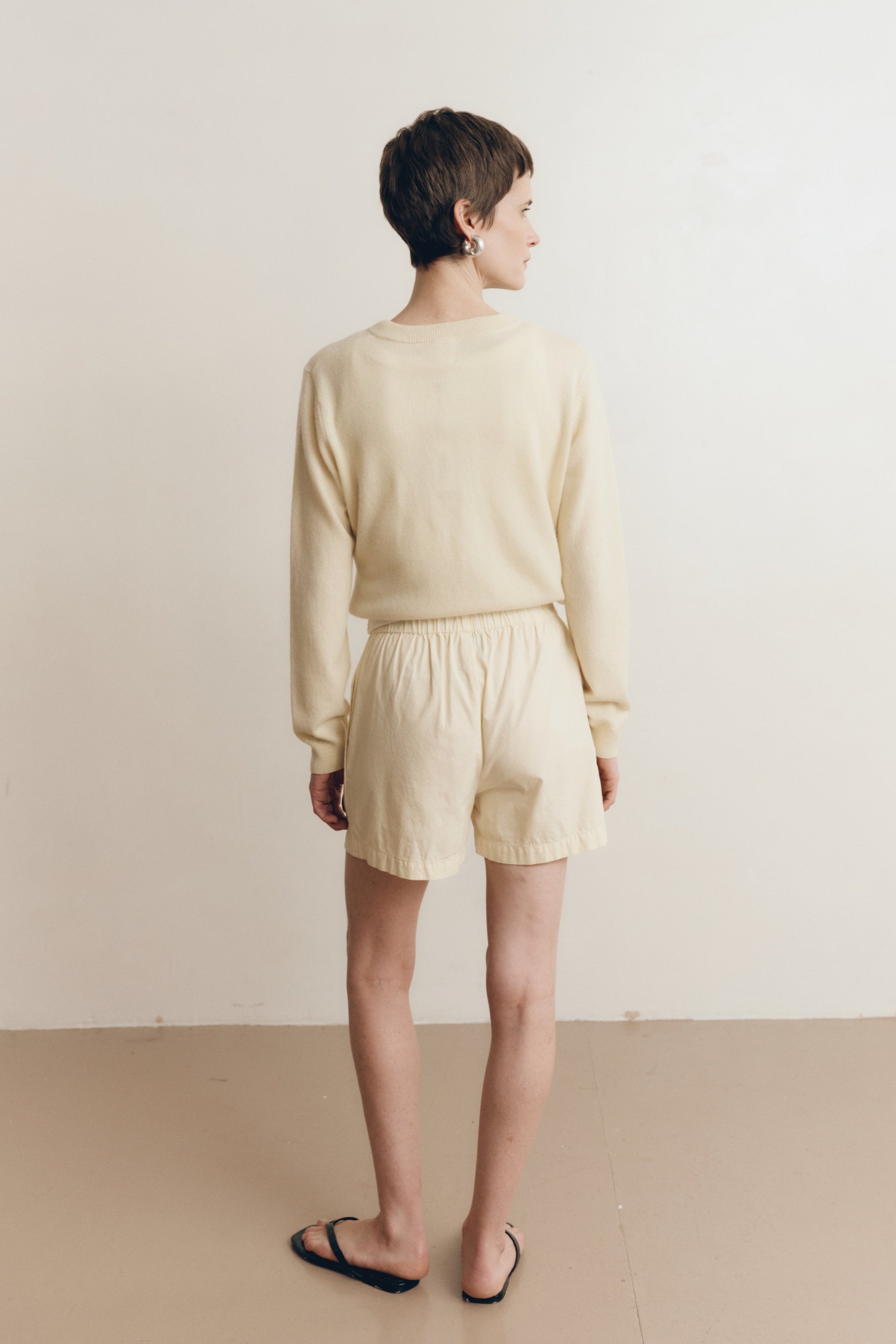Posed female model wearing 100% cotton poplin relaxed shorts with a button closure  in pale yellow paired with a pale yellow matching cashmere cardigan and black sandals - VIR VITA
#color_butter