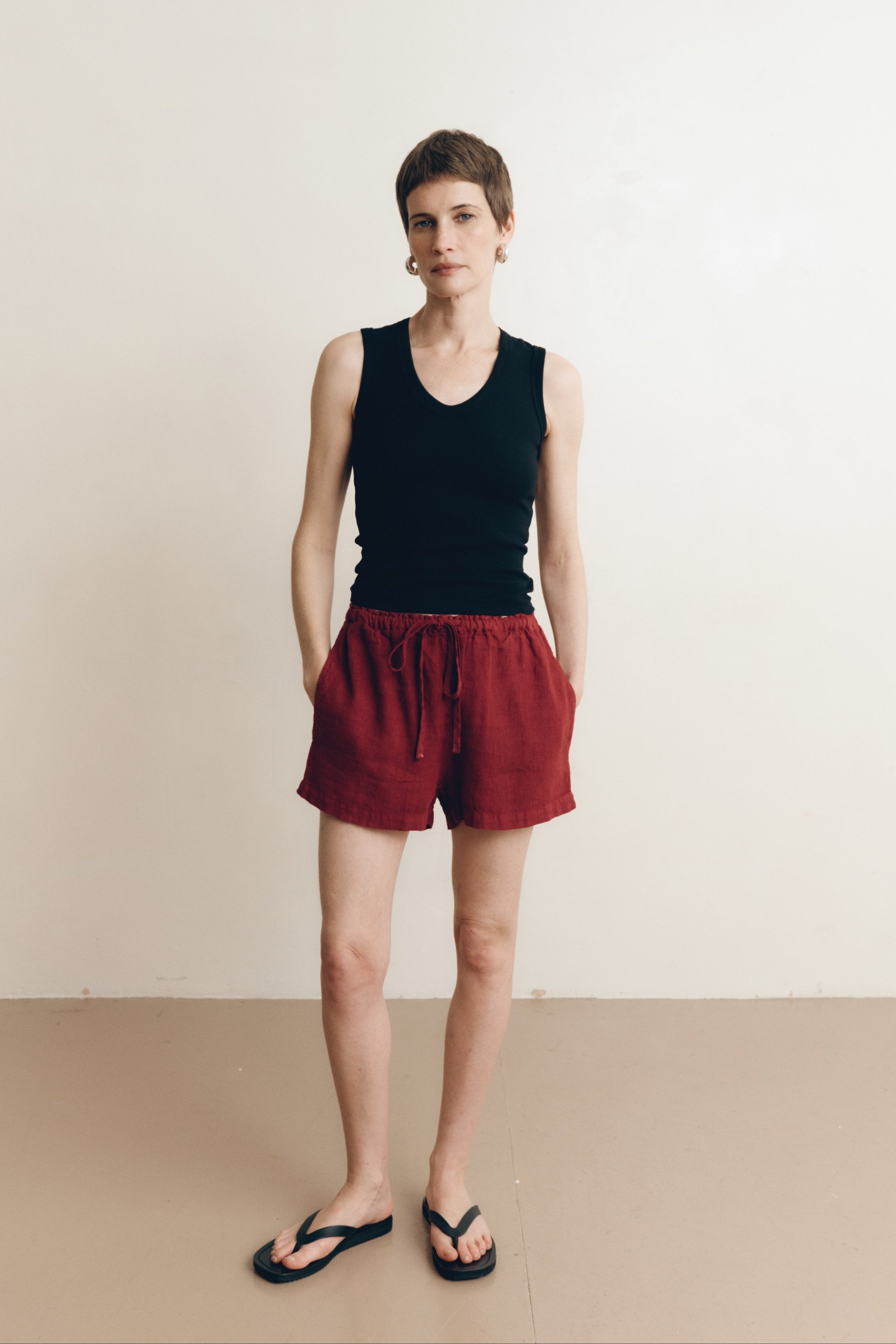 Posed female model wearing sustainable 100% linen drawstring shorts in dark red paired with a black v-neck tank top, and black sandals - VIR VITA
#color_sun dried tomato