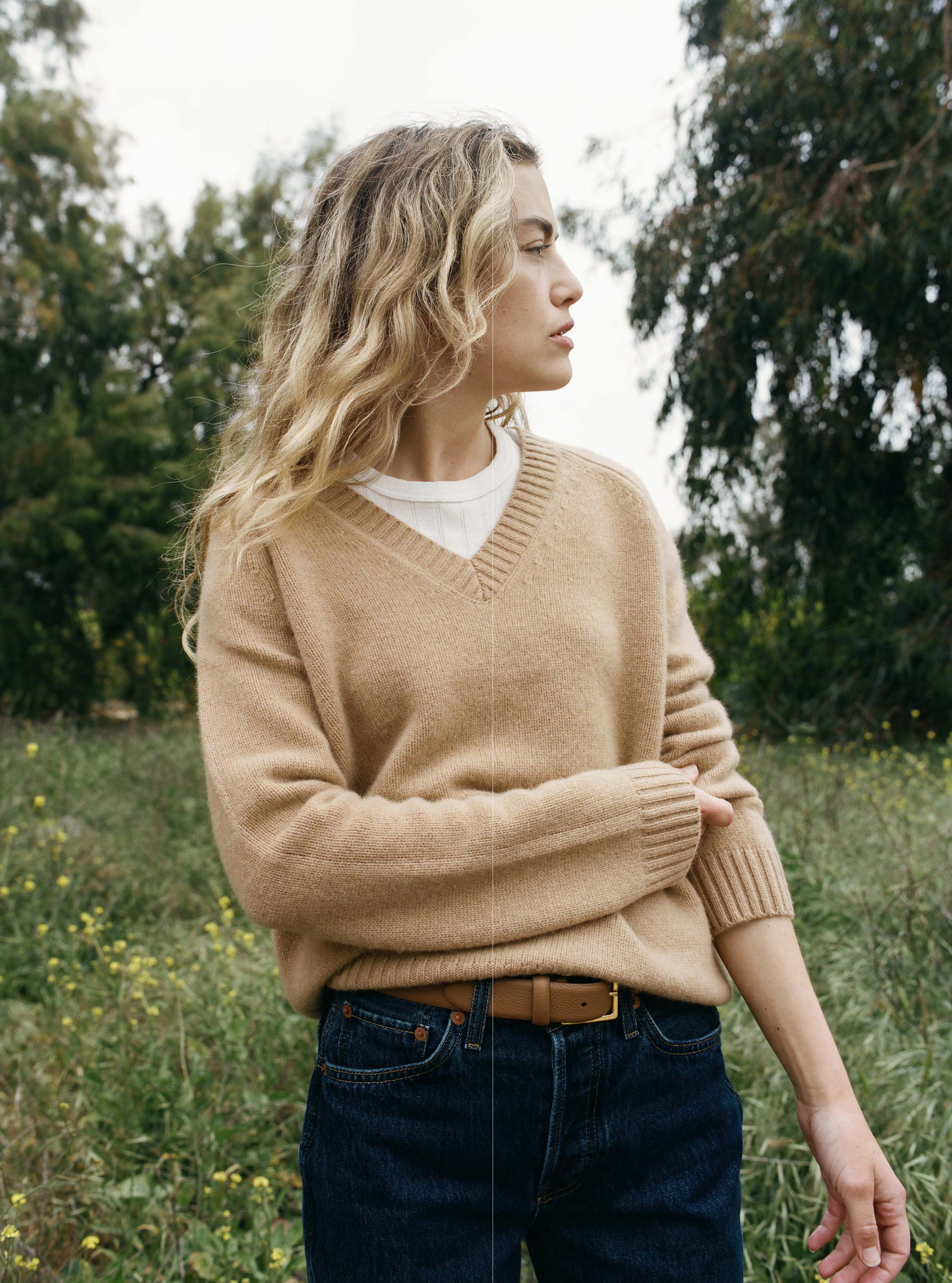 Female model standing in a field wearing an ultra-soft luxurious cashmere pullover sweater featuring a v-neck design in a light brown color paired with dark blue jeans, a brown belt, and a white pointelle tee - VIR VITA
#color_desert
