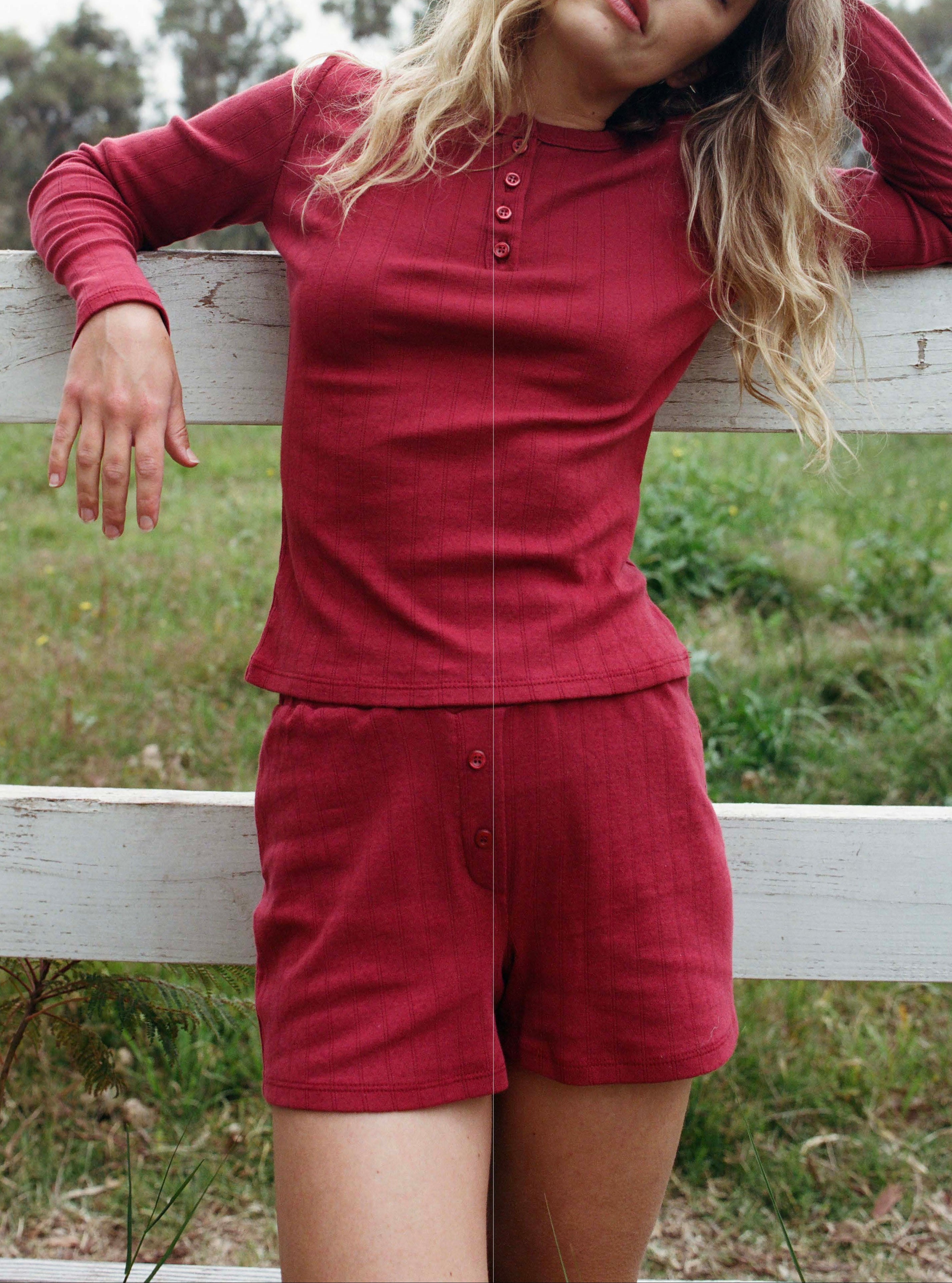 Female model leaning against a fence wearing a dark red cotton pointelle henley long sleeve top with matching buttons paired with cotton pointelle lounge shorts in dark red - VIR VITA
#color_sun dried tomato